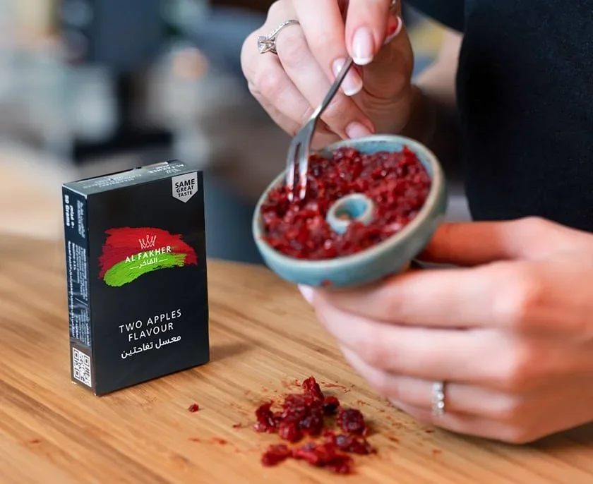 Person filling a shisha head with a spoon, next to a carton of Al Fakher Two Apples flavored shisha tobacco on a wooden table.