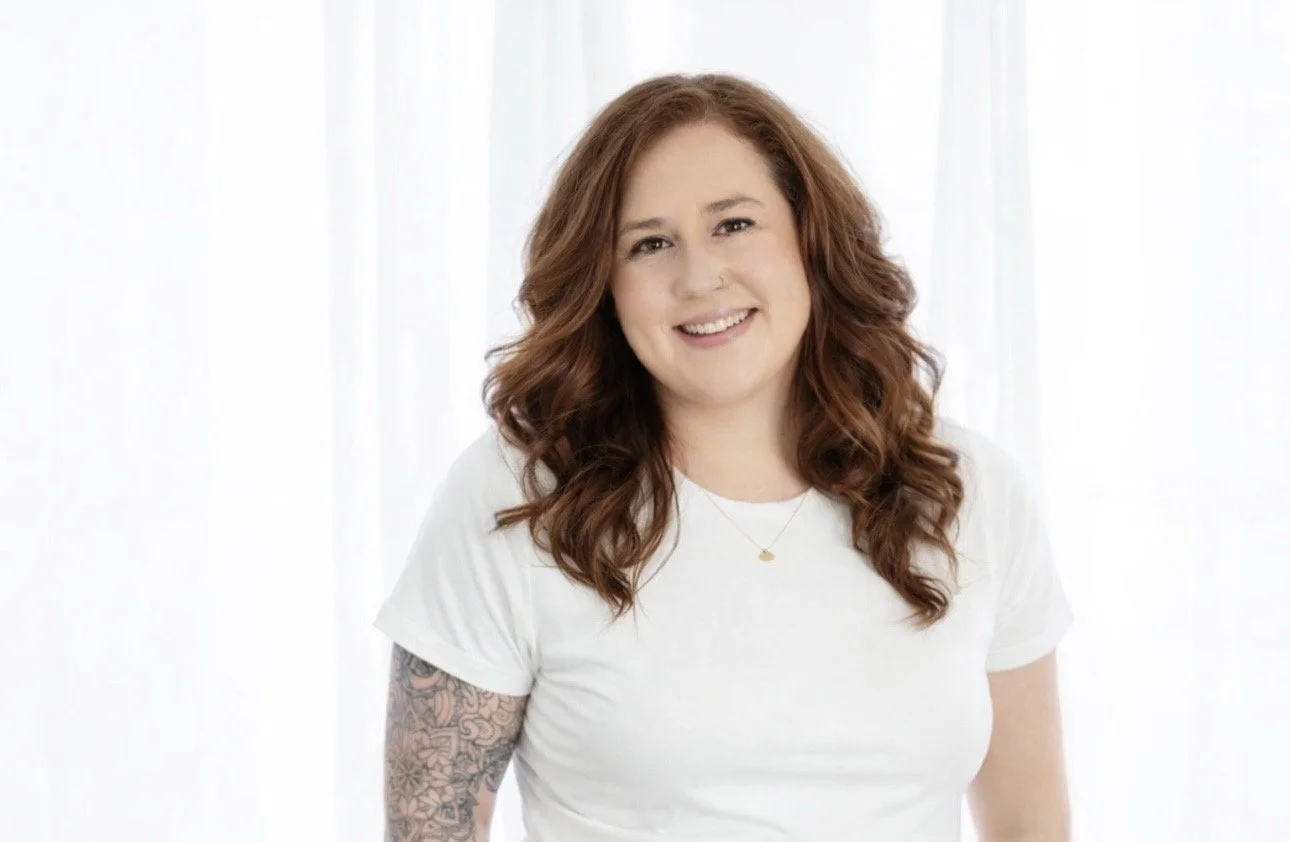 A woman with long, wavy, reddish-brown hair smiling, wearing a white t-shirt and a gold necklace, standing in front of a light-colored curtain background.