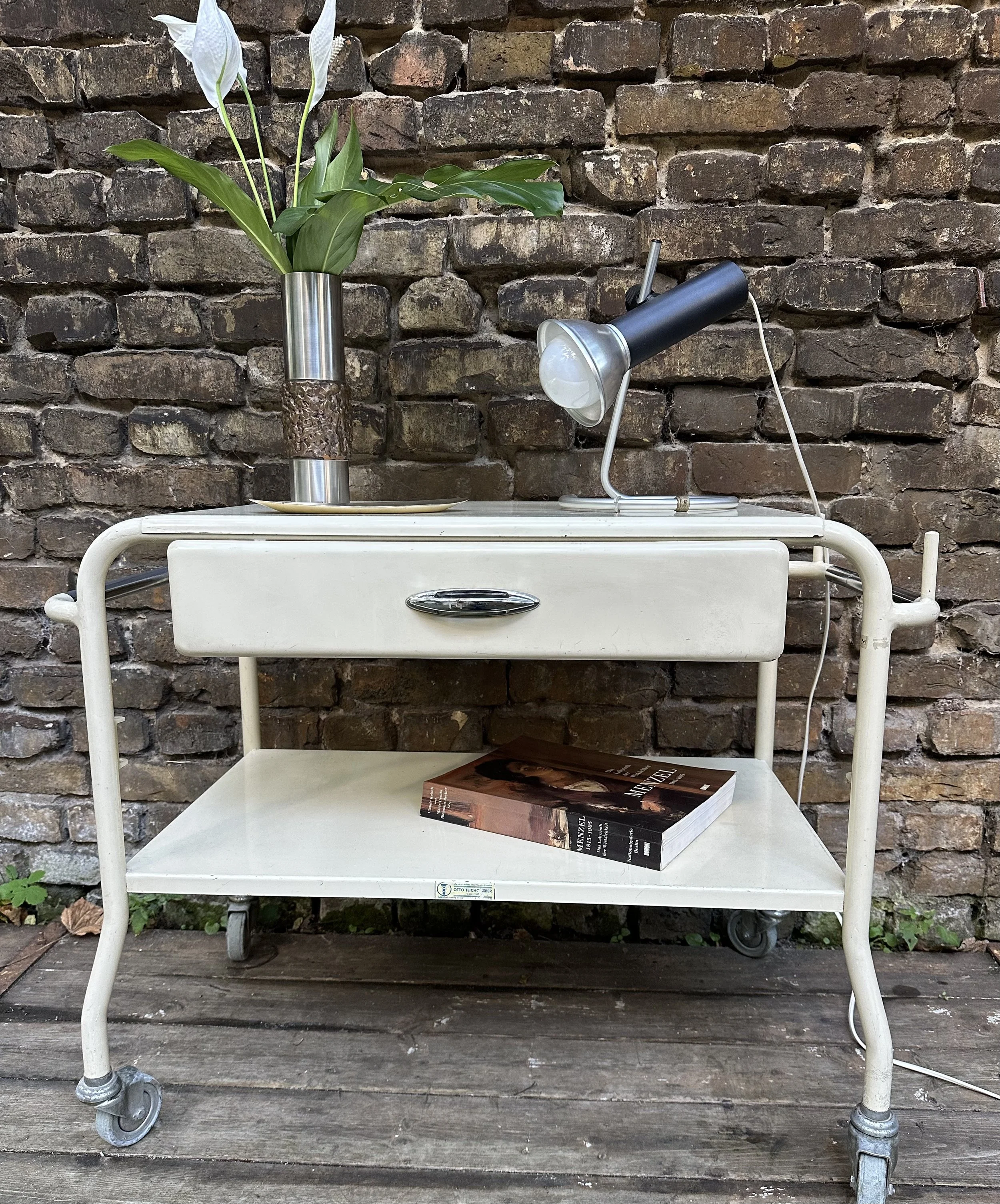 Weißer Vintage Rollwagen mit Lampe und Vase vor Backsteinwand im Store-Umfeld