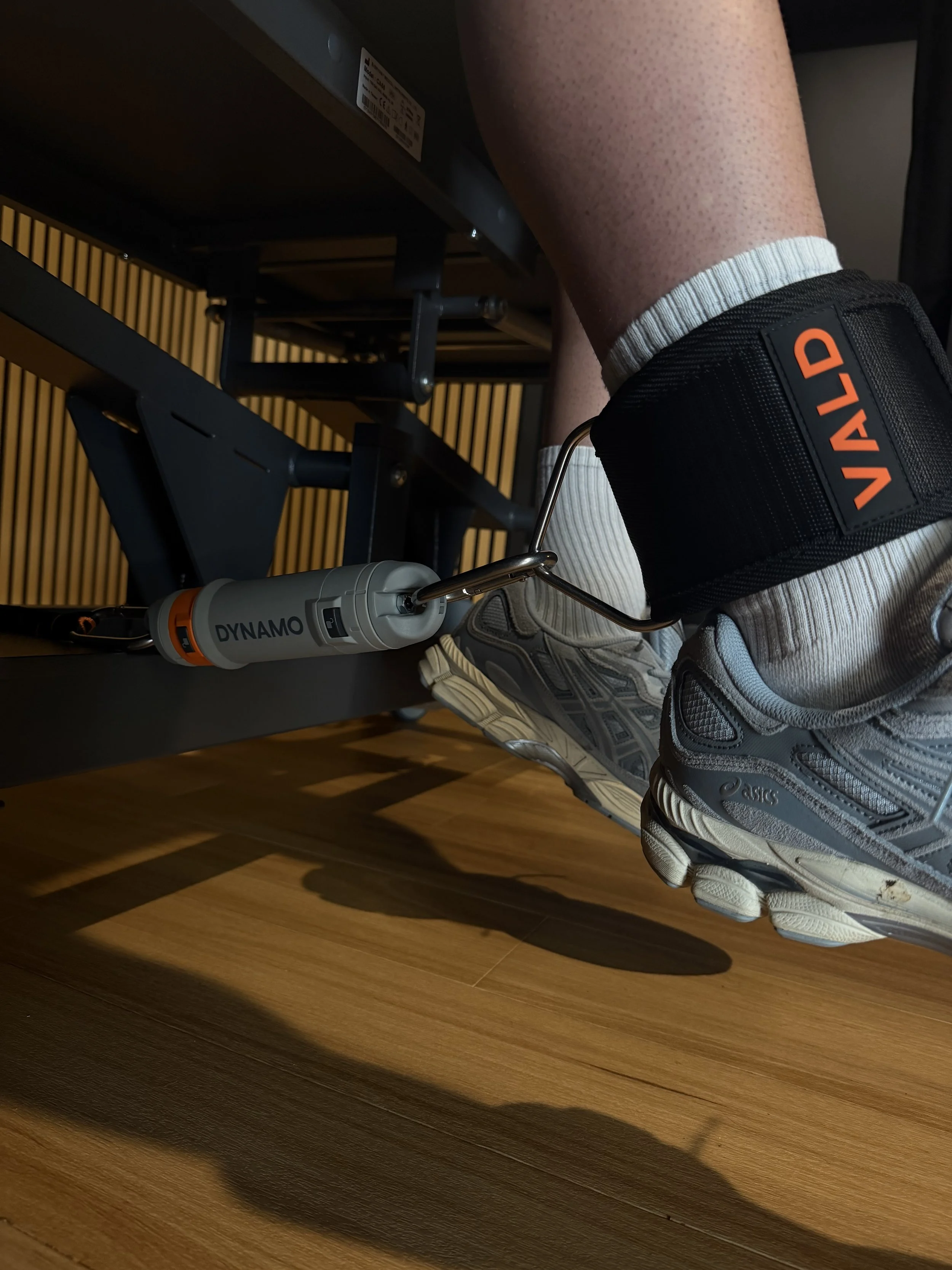 A person wearing gray athletic shoes and white socks with gray stripes is attached to a foot pedal exerciser with a gray and black ankle cuff labeled 'VALD.' The exerciser has a label 'DYNAMO' and is placed on a wooden floor.