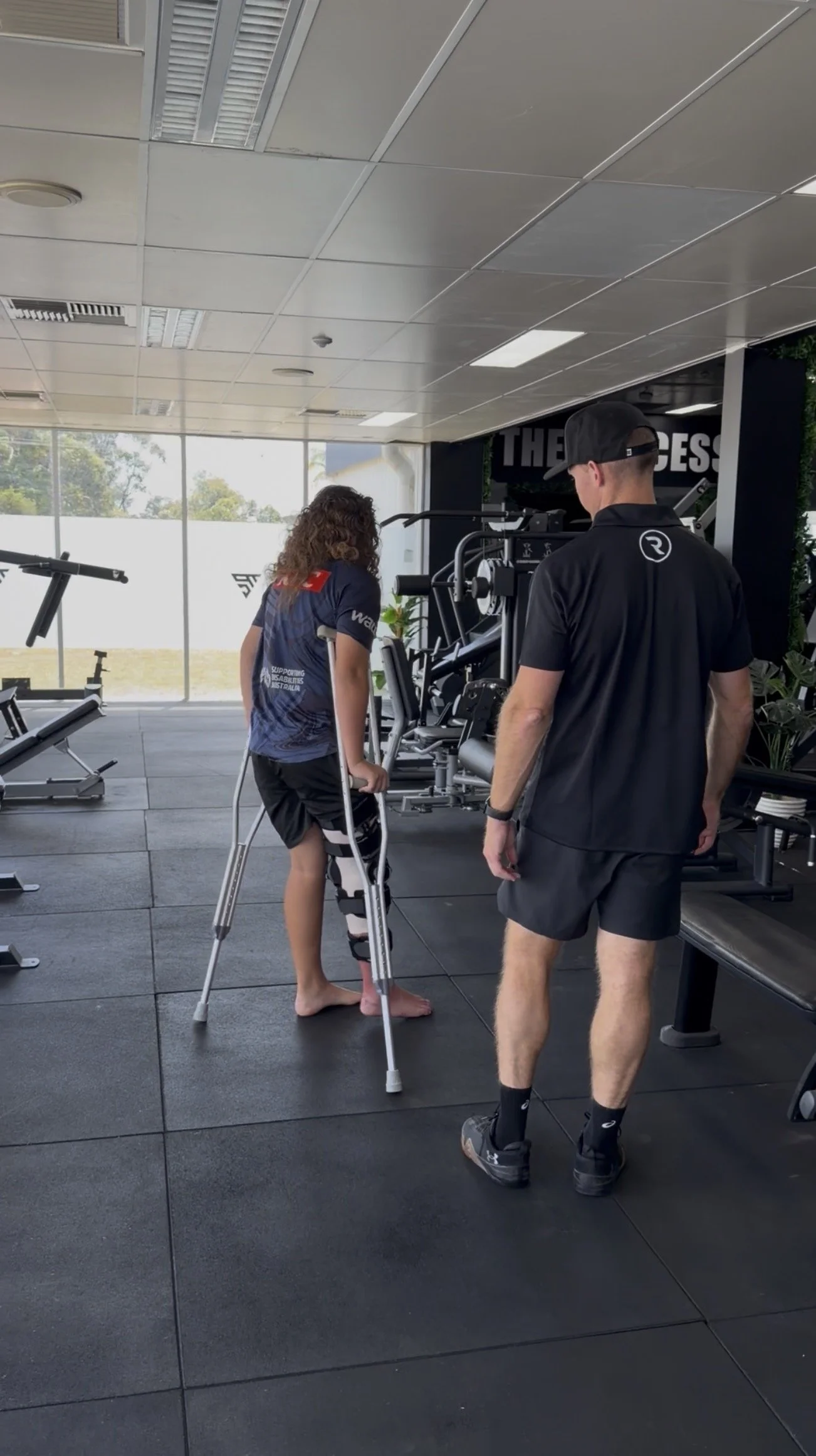 A woman with a leg injury using crutches in a gym while talking to a trainer.