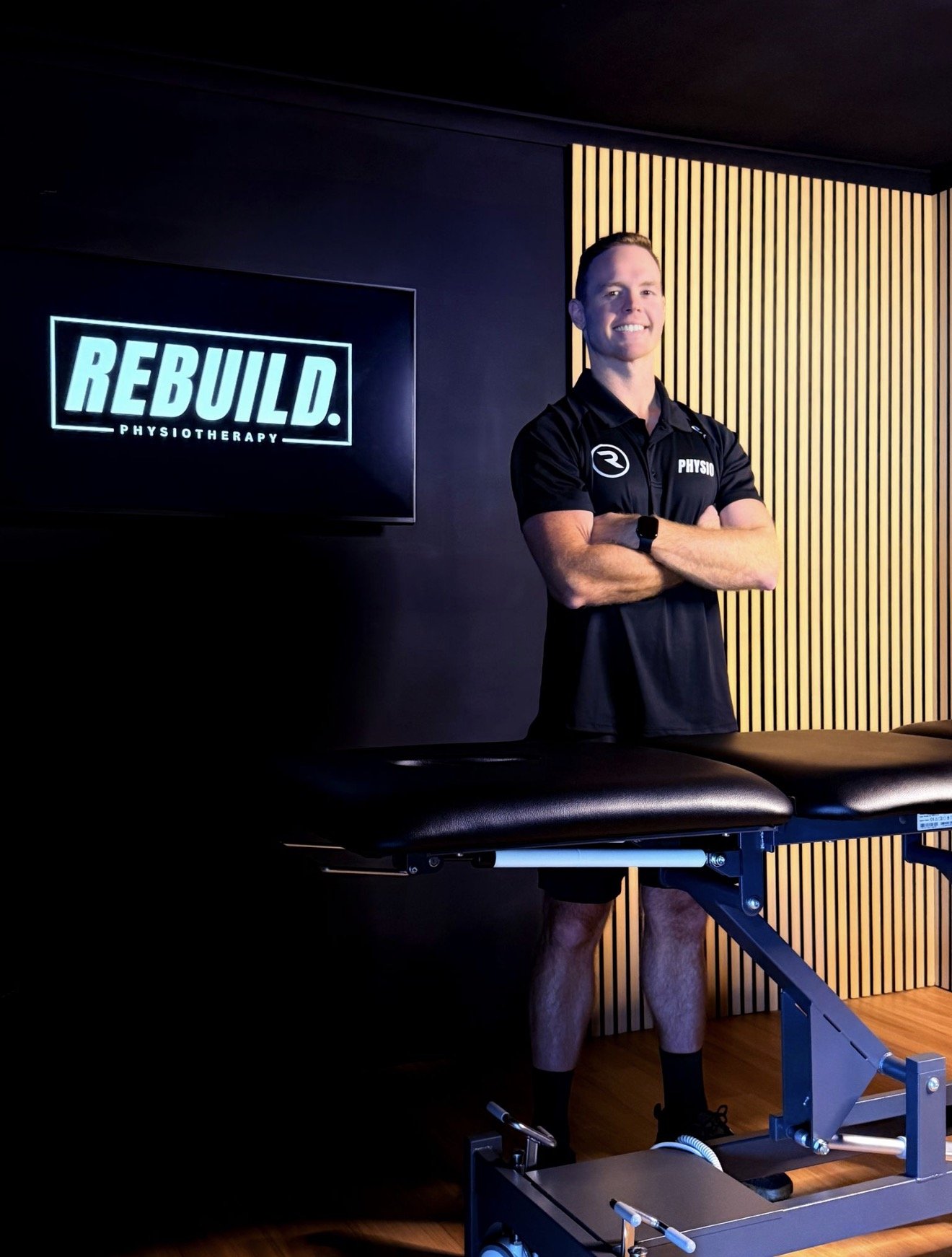 A man in a black sports shirt and shorts standing with arms crossed in a physiotherapy clinic, smiling. There is a wall-mounted tv screen with the logo 'REBUILD. PHYSIOTHERAPY' and a treatment table in front.