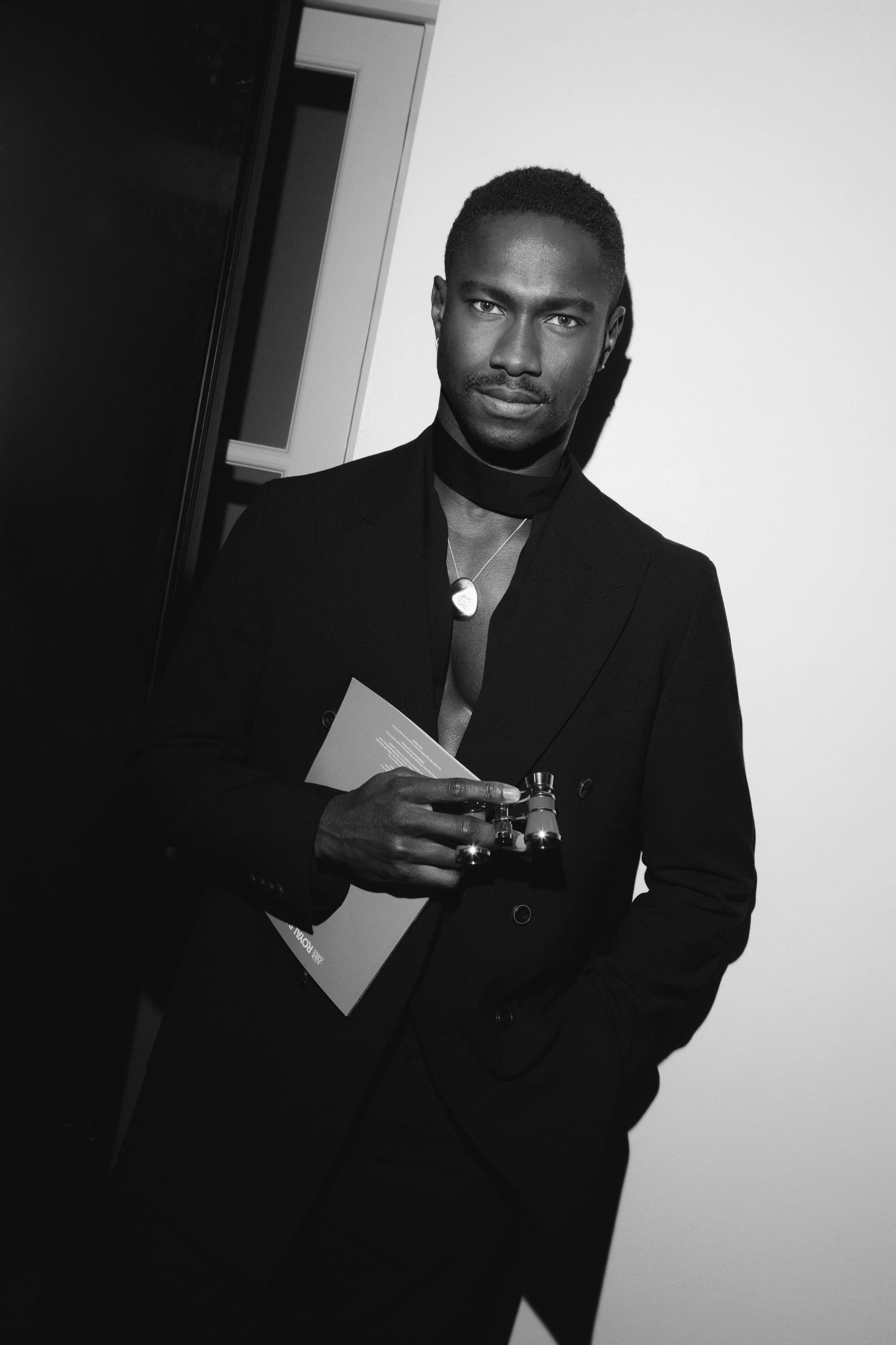 A black-and-white photo of a man in formal attire holding a brochure and possibly a small bottle of perfume, standing against a plain background.