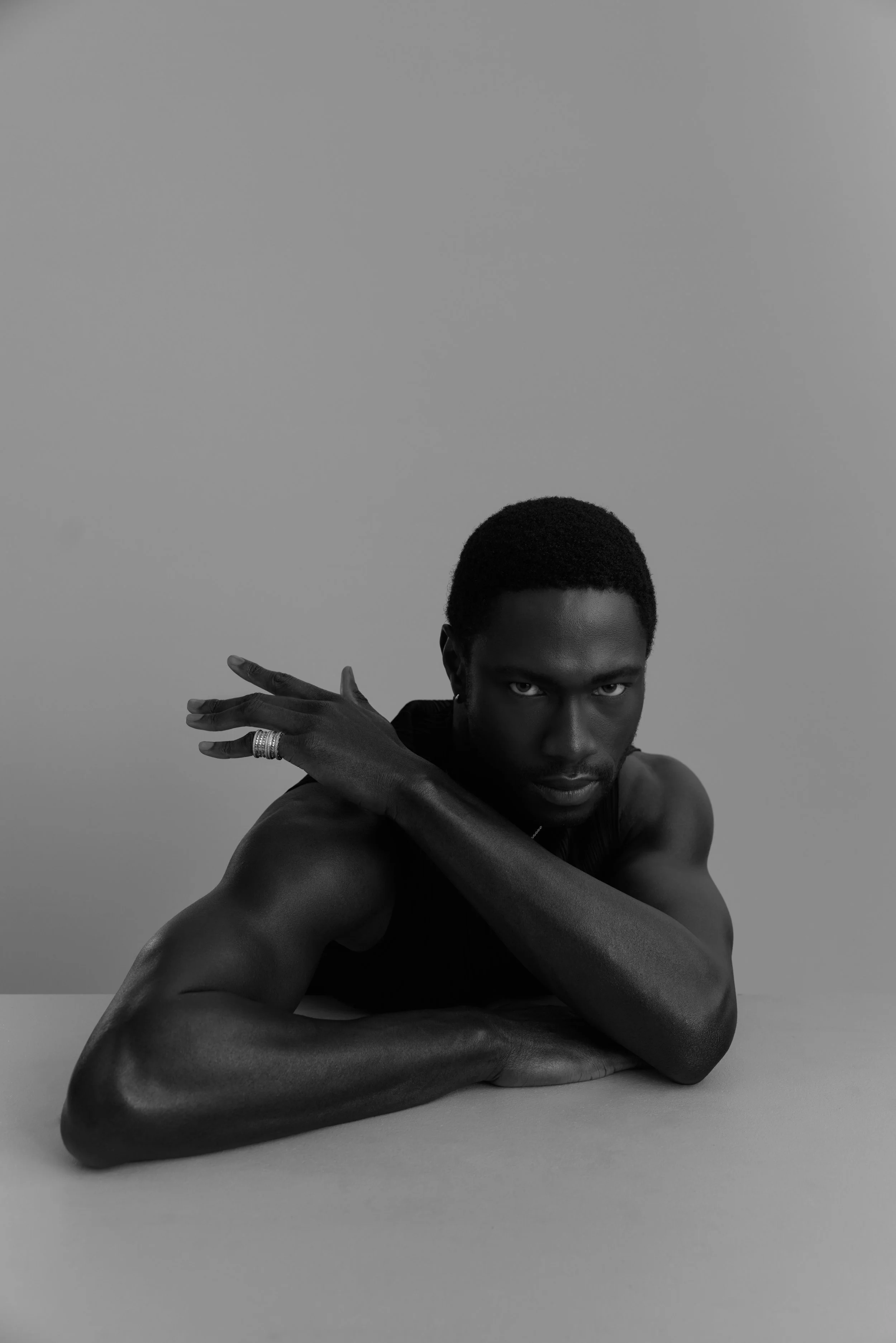 A black and white portrait of a shirtless man with dark skin, leaning on his forearms on a flat surface, with one arm crossed over the other, looking directly at the camera with intense expression.