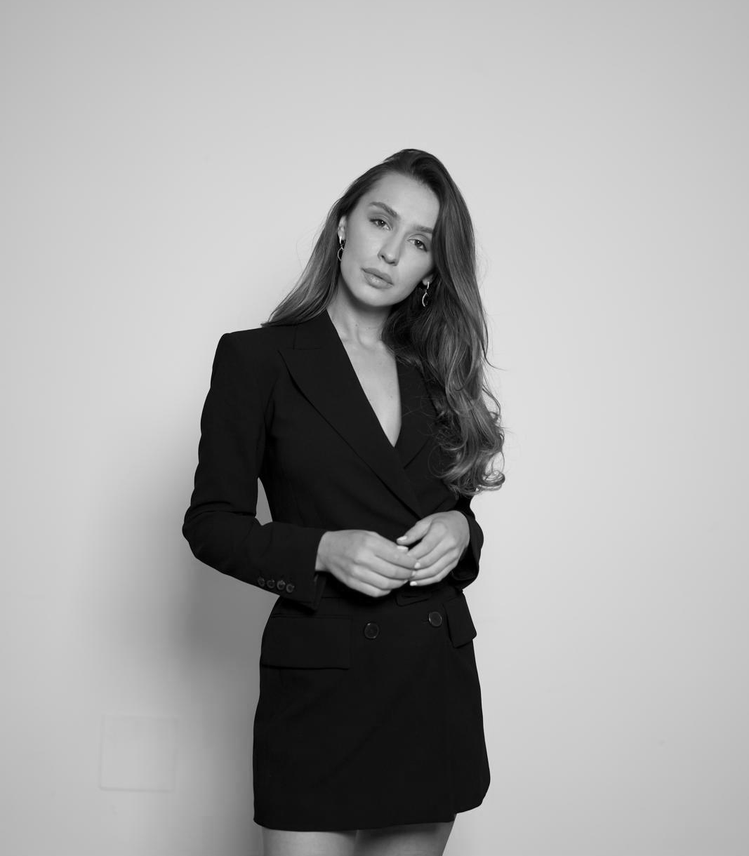 Black and white portrait of a woman in a formal black blazer and skirt, standing against a plain background.