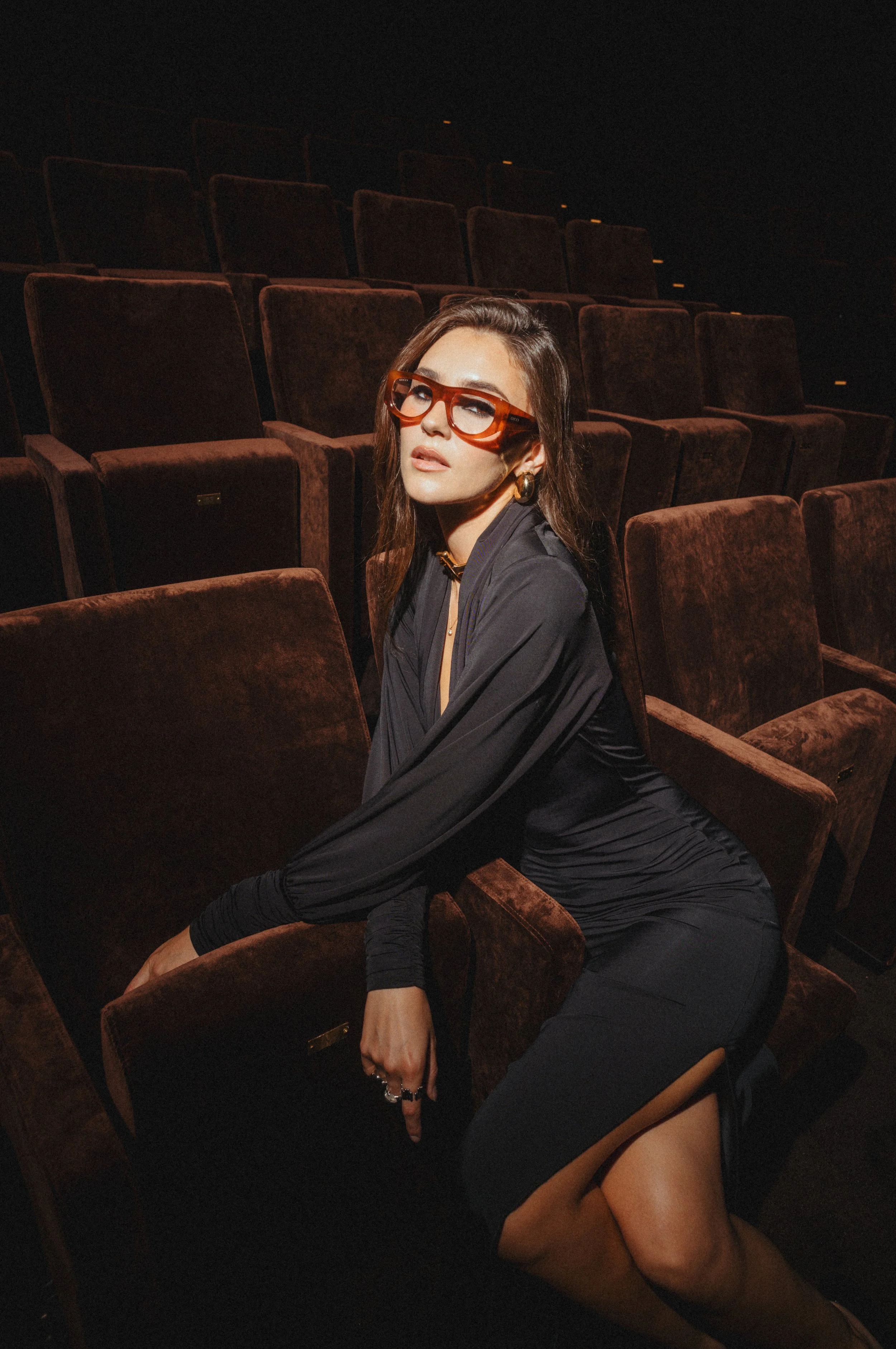 A woman sitting in a dimly lit theater with brown velvet seats, wearing orange sunglasses and a black dress.