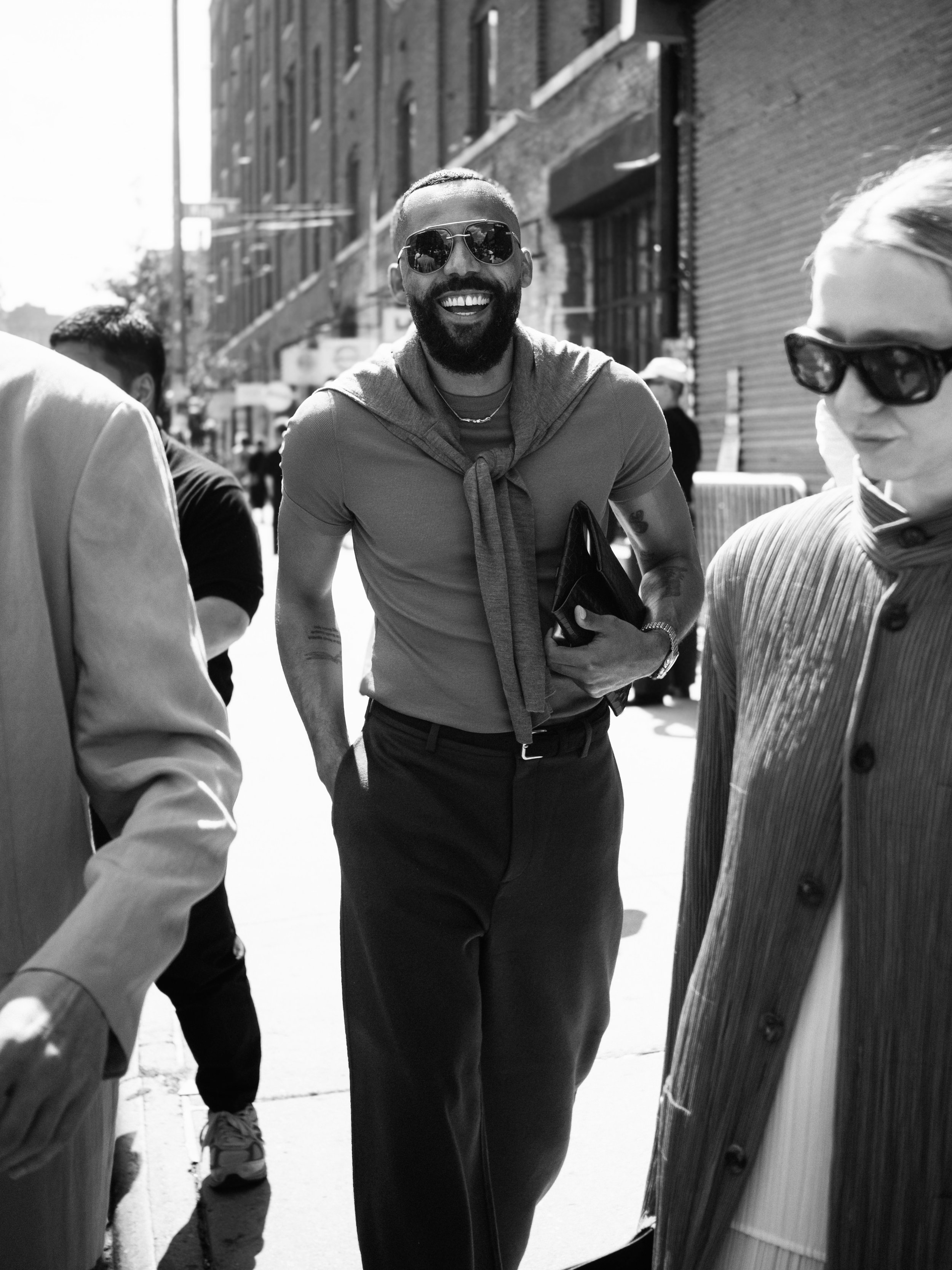A smiling man with a beard and sunglasses walking outdoors in an urban area, holding a small black bag, surrounded by other people.