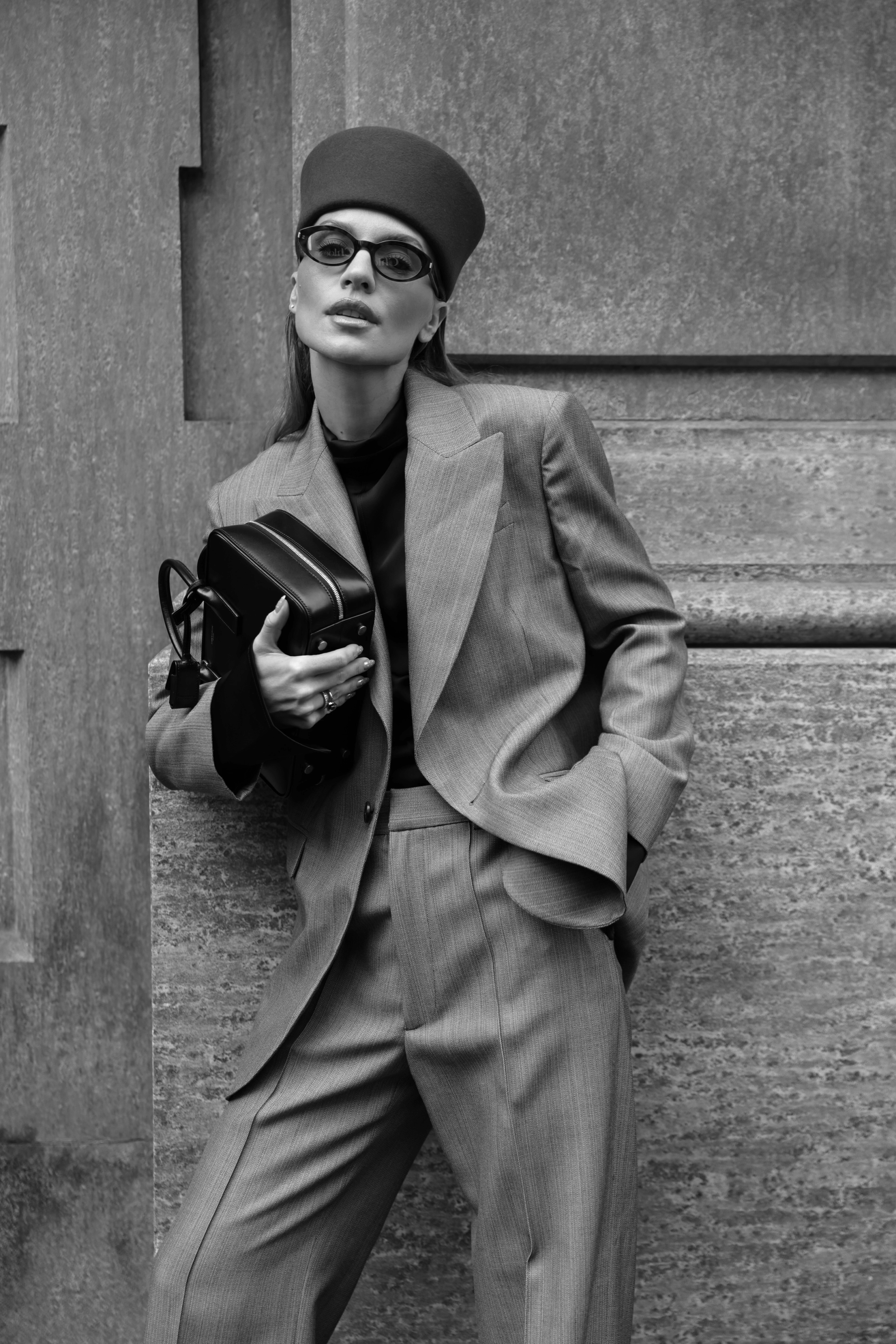 A woman in a tailored suit, wearing sunglasses and a hat, leaning against a textured wall, holding a small handbag.