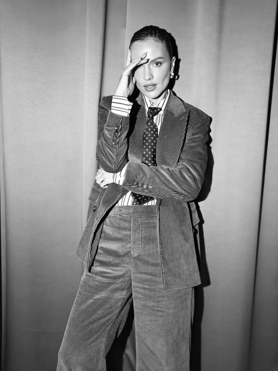 Black and white photo of a woman wearing a pinstripe suit, standing with one arm crossed and the other hand touching her forehead.