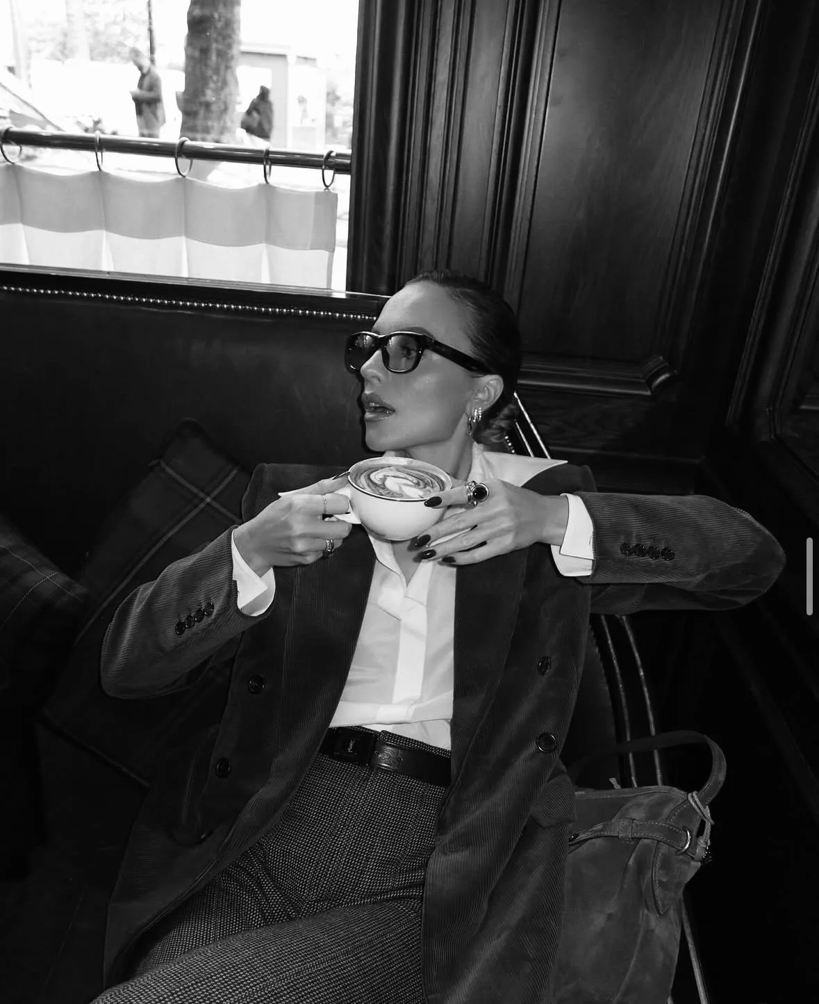 A woman with glasses and earrings is seated in a cozy cafe, holding a cup of coffee, dressed in a blazer, white shirt, and patterned pants, with a bag on the seat beside her.