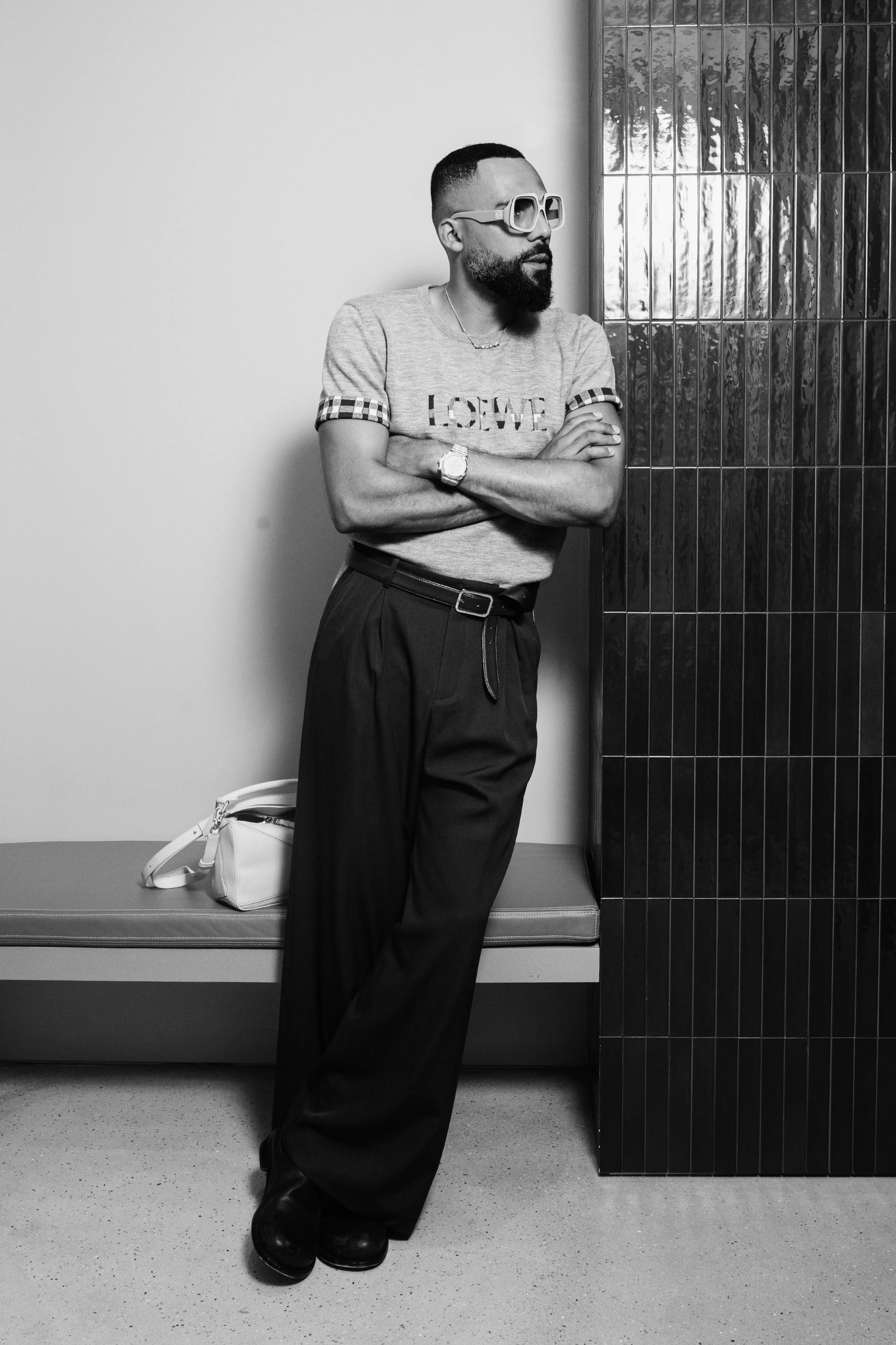 A man with a beard and glasses standing against a wall with arms crossed, wearing a Loewe t-shirt, black wide-leg trousers, and a watch, in a modern indoor setting.