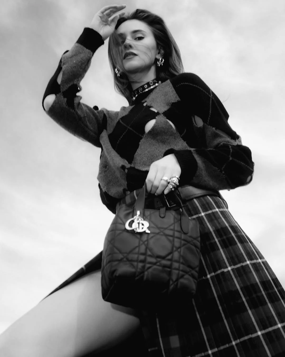 A woman posing outdoors, wearing a patterned sweater, plaid skirt, and jewelry, holding a quilted purse with a Dior charm.