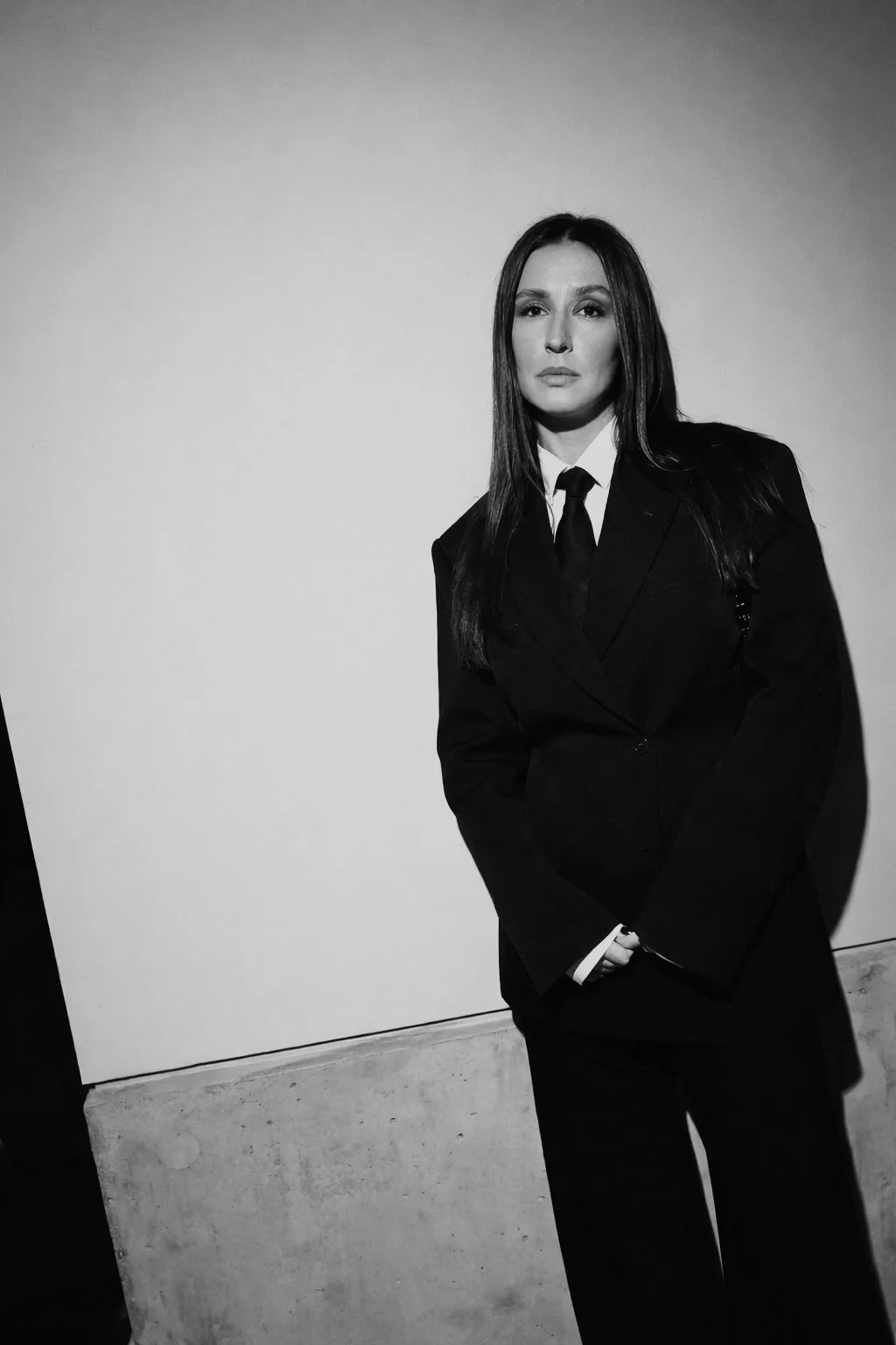 A black and white photo of a woman in a business suit standing against a plain wall, looking at the camera with a serious expression.