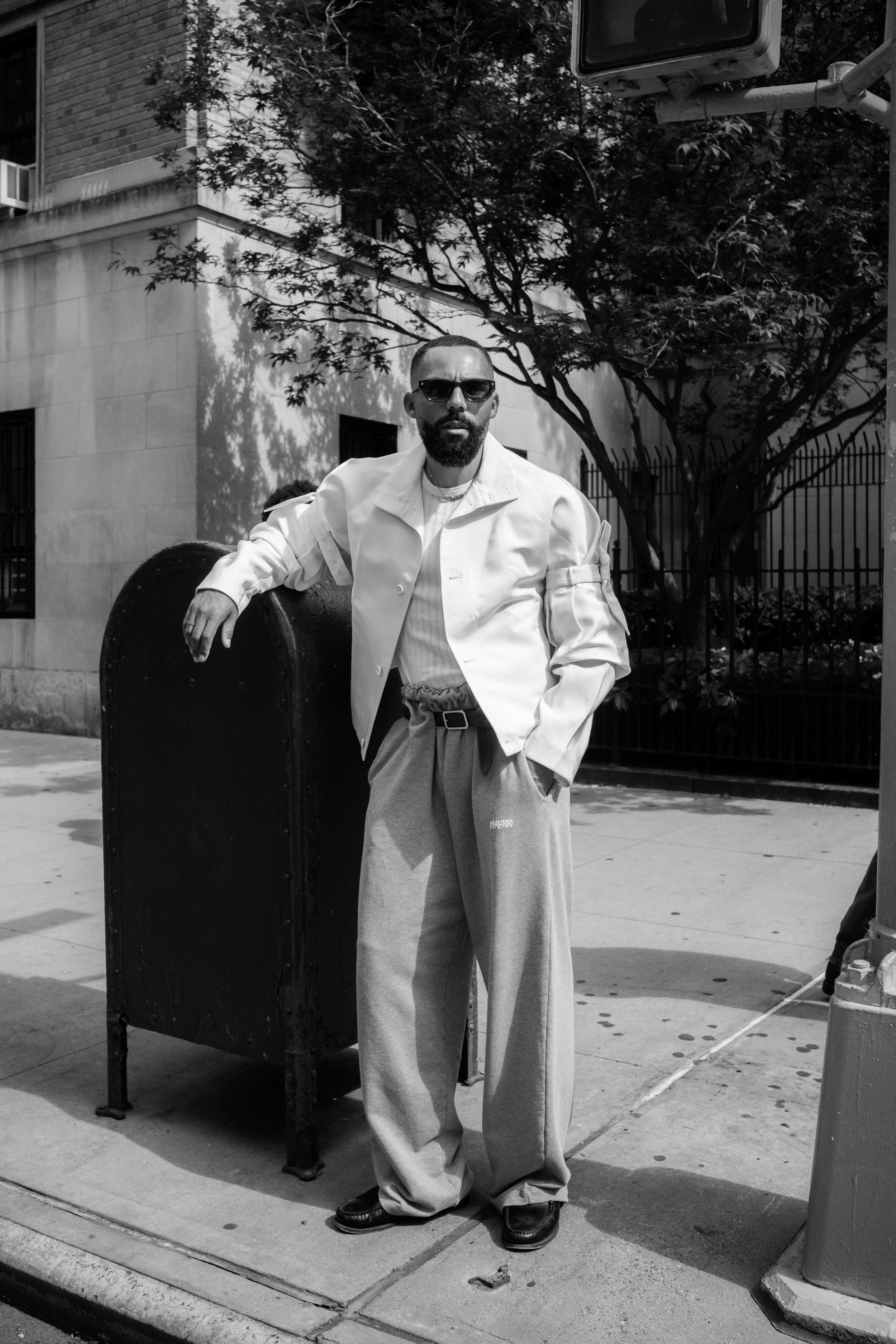A man with a beard wearing sunglasses, a white jacket, and wide pants standing next to a mailbox on a city sidewalk, with trees and a building in the background.