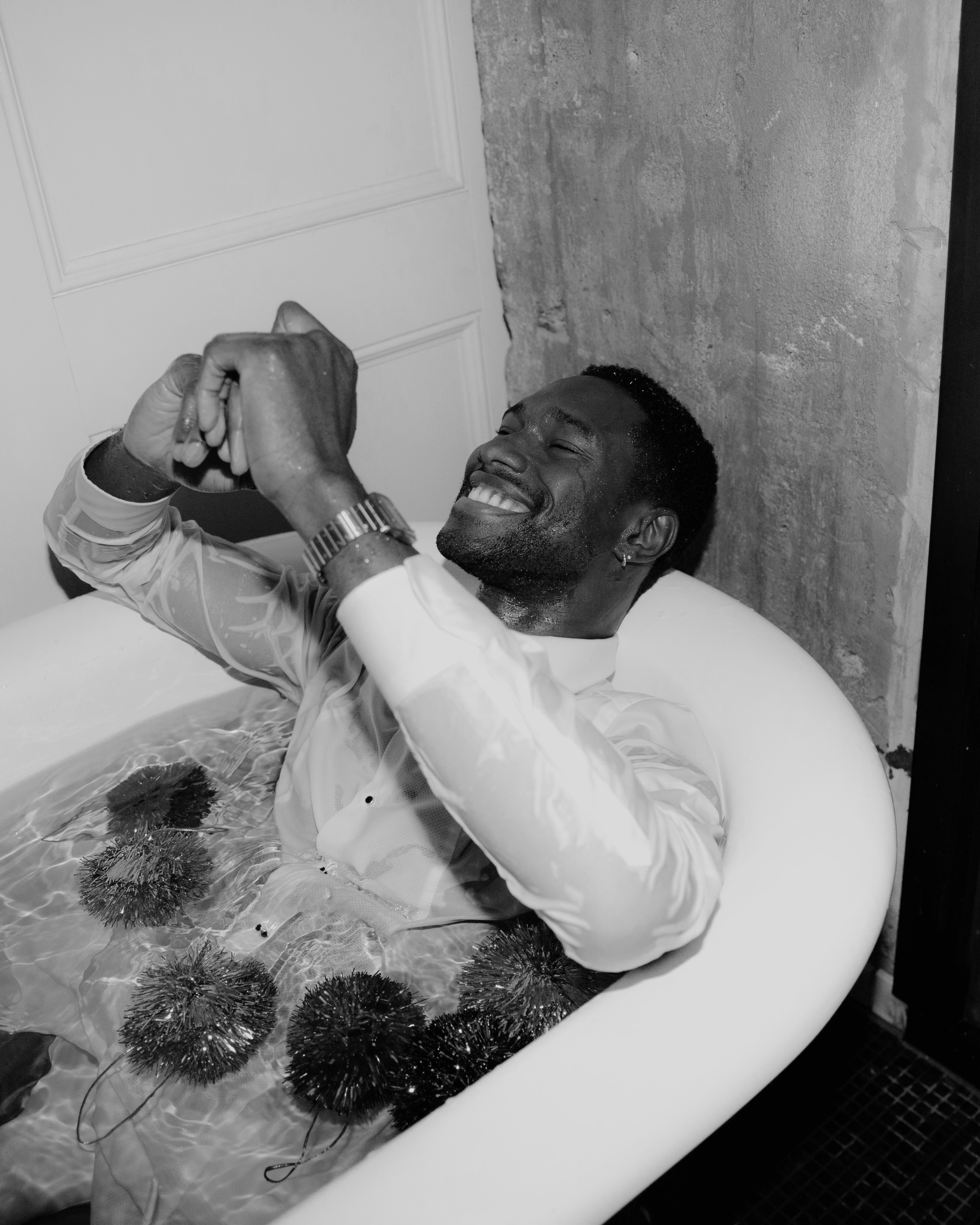 A man smiling and relaxing in a bathtub filled with water and submerged spiky balls, wearing a shiny long-sleeve shirt, with a textured wall and door in the background.