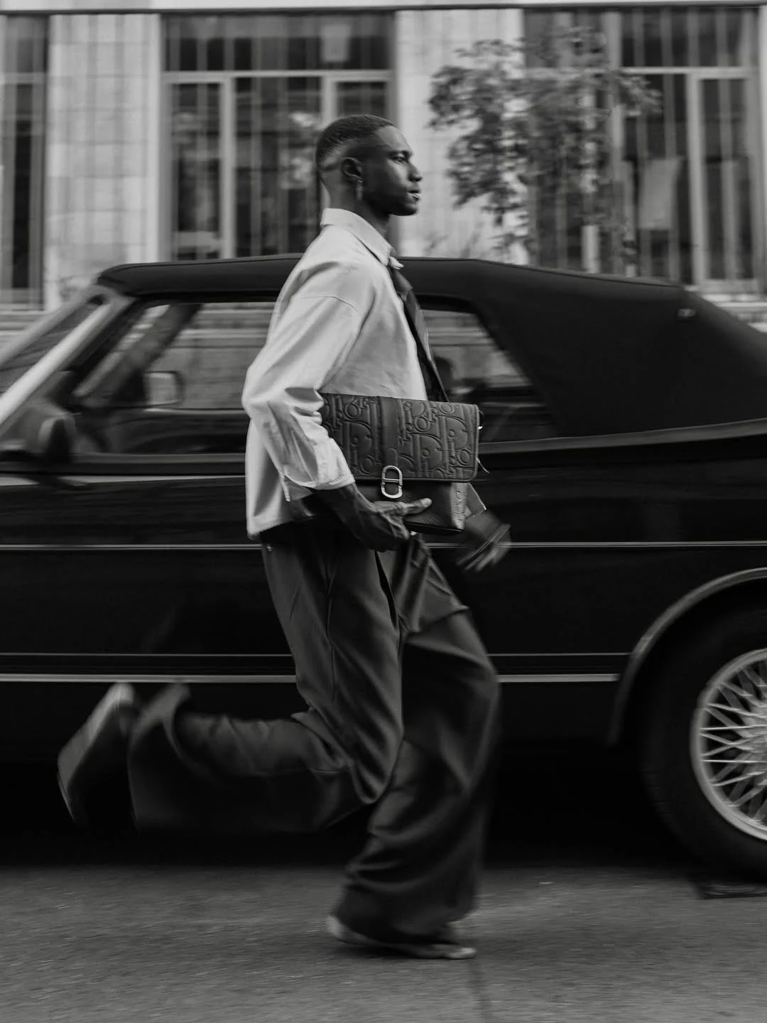 A person walking briskly in front of a moving black car on a city street, holding a designer bag, in black and white.
