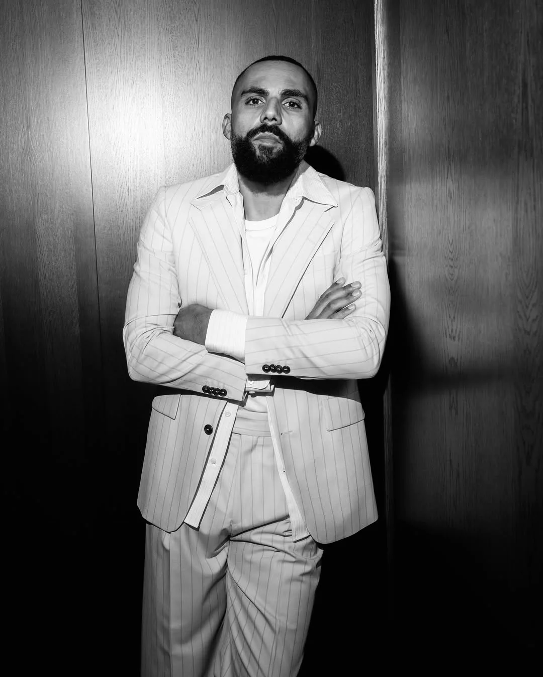 A man with a beard in a white pinstripe suit standing against a wooden wall with arms crossed.