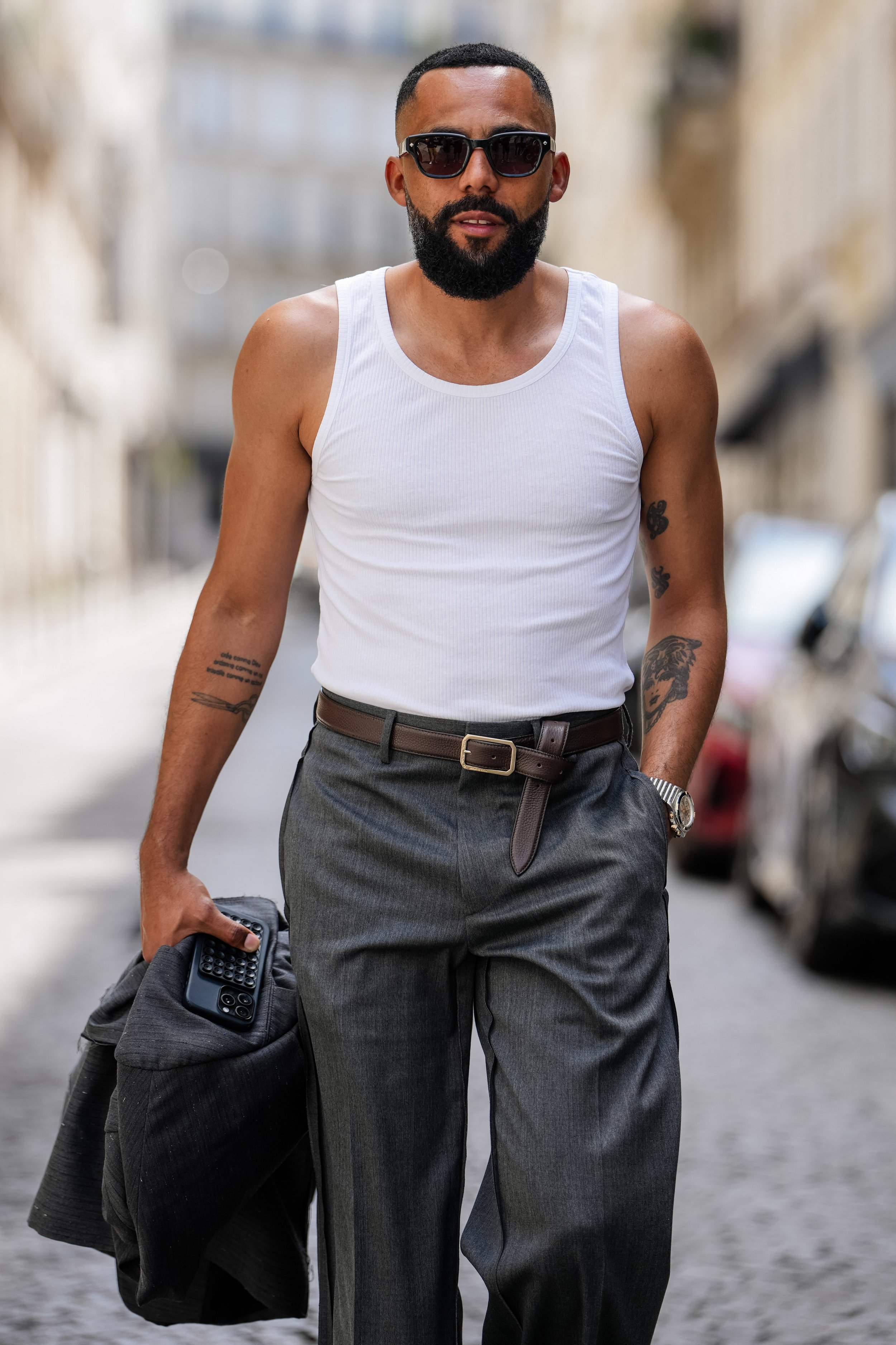 A stylish man with a beard and sunglasses walking on a city street, wearing a white tank top and gray pants, holding a jacket and a phone.