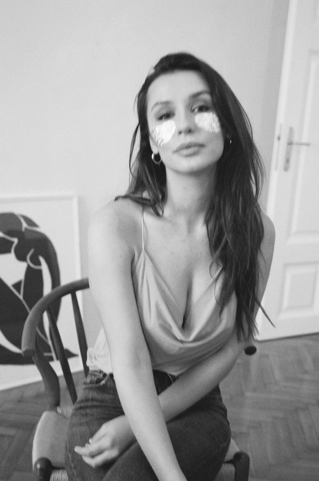 Black and white photograph of a woman with long dark hair sitting on a wooden chair, with her arms crossed in her lap. She is wearing a sleeveless top with a deep neckline and earrings, and has aluminum foil on her cheeks.