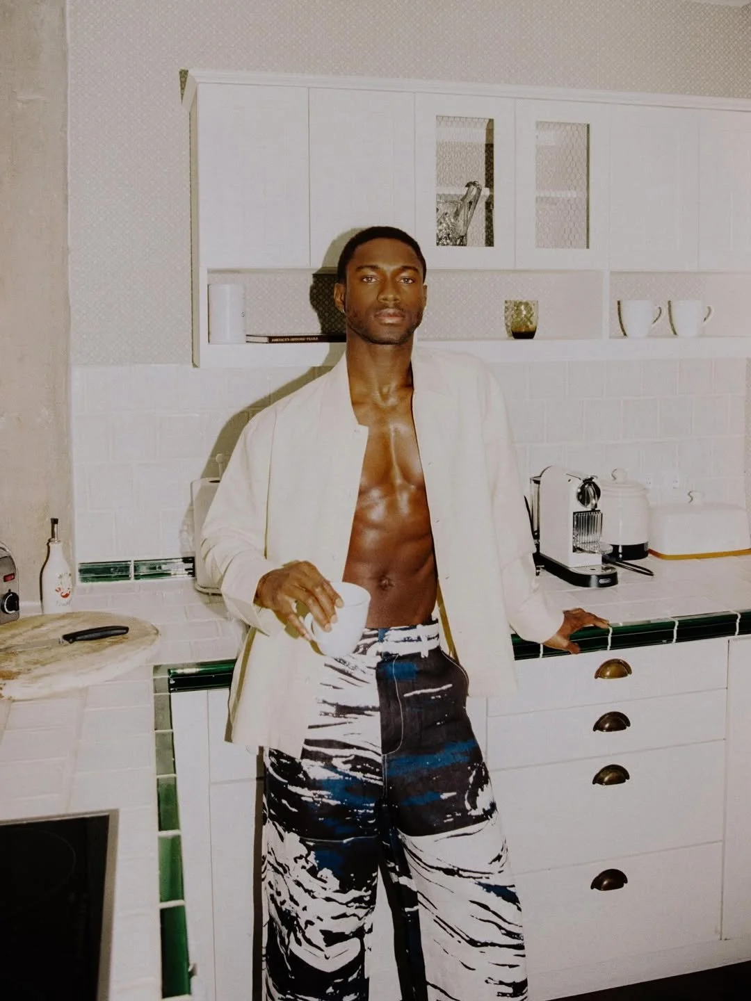 A shirtless man with a white blazer and patterned pants holding a white mug standing in a white kitchen.