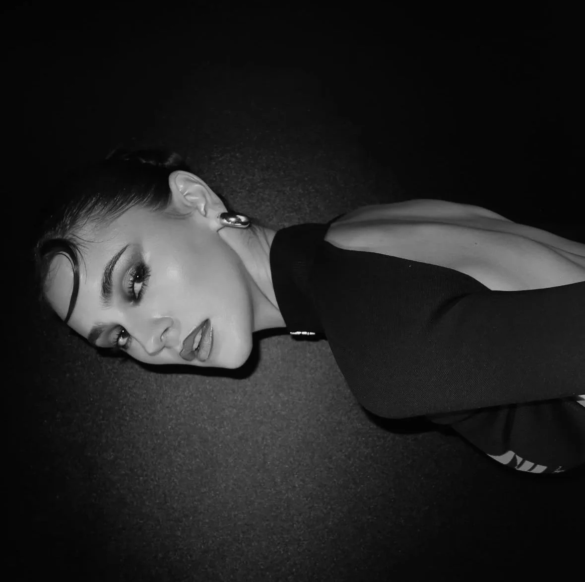 Black and white photo of a woman with dark hair, makeup, and earrings lying on a dark surface, looking at the camera.