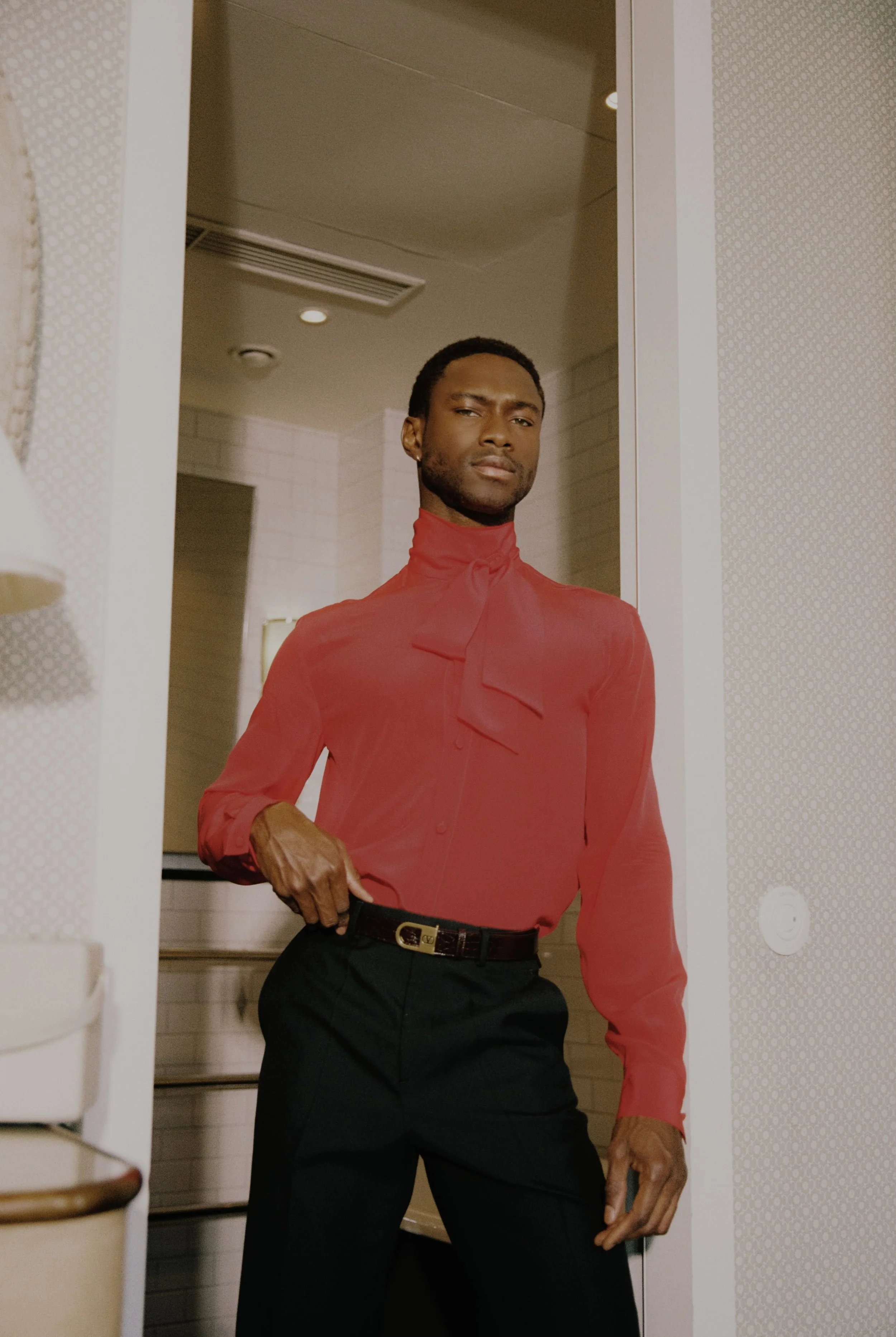 Man standing and looking at the camera, wearing a red blouse with a bow and black pants, in an indoor setting.