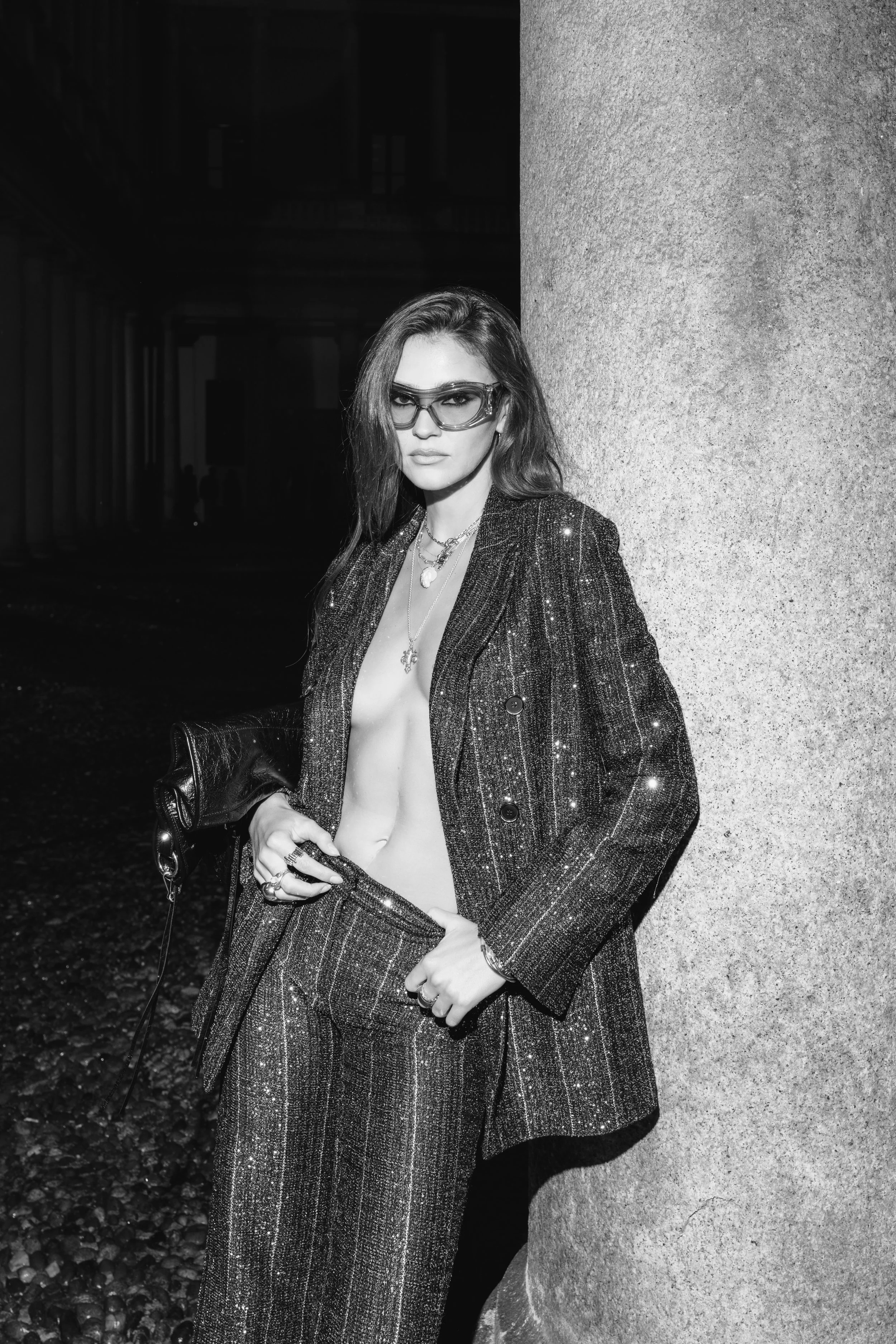 A woman wearing sunglasses and a sparkly suit with no top underneath, standing against a stone wall, holding a purse in her right hand, outdoors at night.