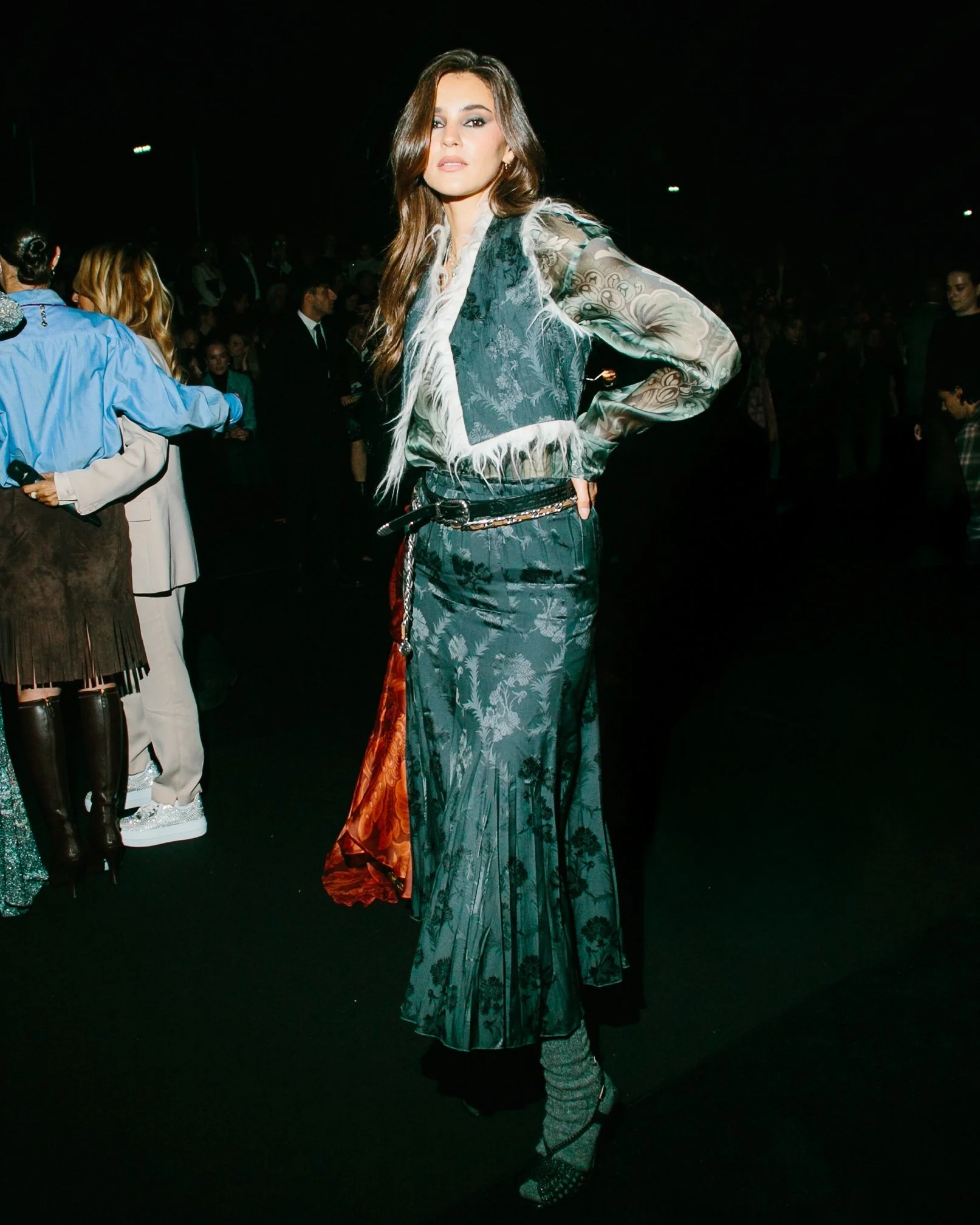 A woman with long brown hair wearing a patterned outfit, standing with one hand on her hip at an event with people in the background.