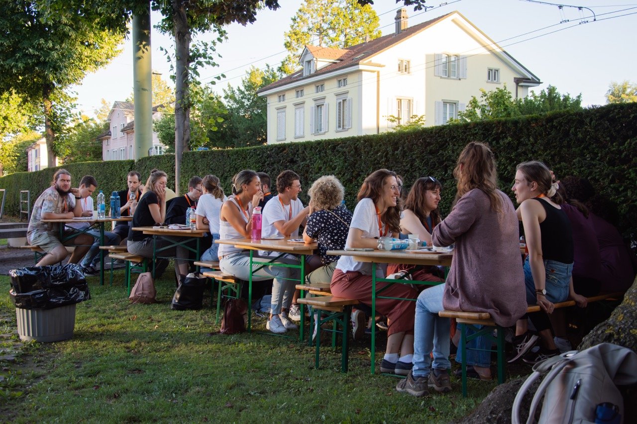 Eine Gruppe von Menschen sitzt an Tischen im Freien, vermutlich bei einem Treffen oder einer Veranstaltung, umgeben von Bäumen und einer Hecke, mit Wohnhäusern im Hintergrund.