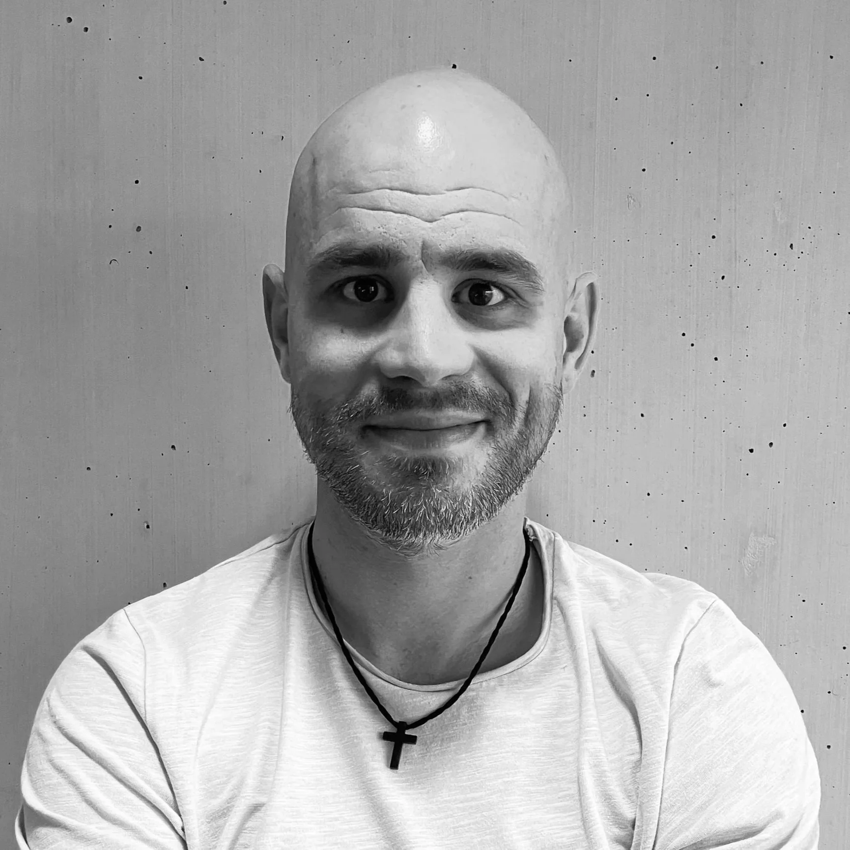 Schwarz-weiß-Fotografie eines lachenden Mannes mit Glatze und Bart, trägt ein T-Shirt und einen Kreuz-Halskette, vor einer schlichten Wand.