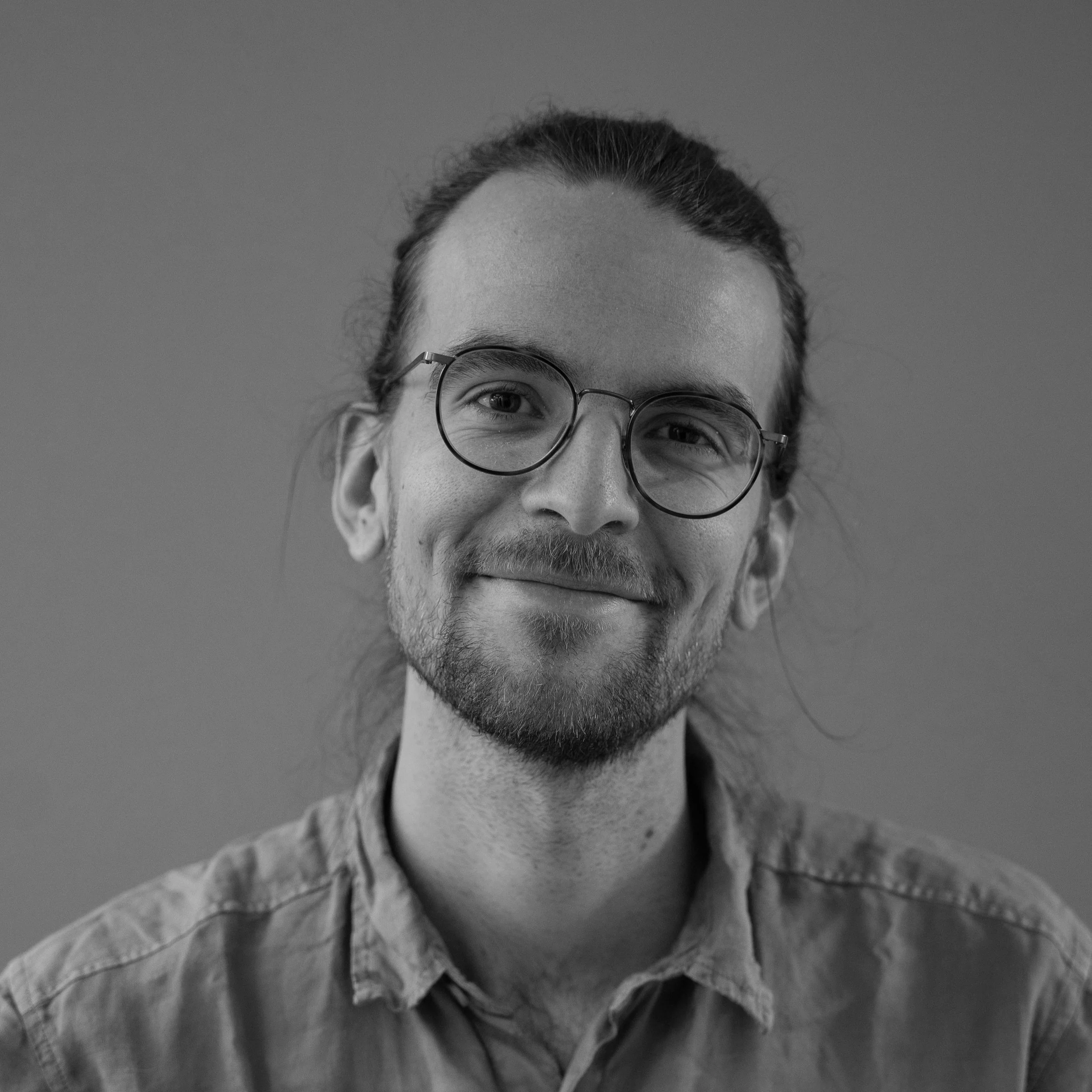 Schwarz-Weiß-Foto eines lächelnden Mannes mit Brille und langem Haar, trägt ein Hemd, vor schlichter Hintergrund.