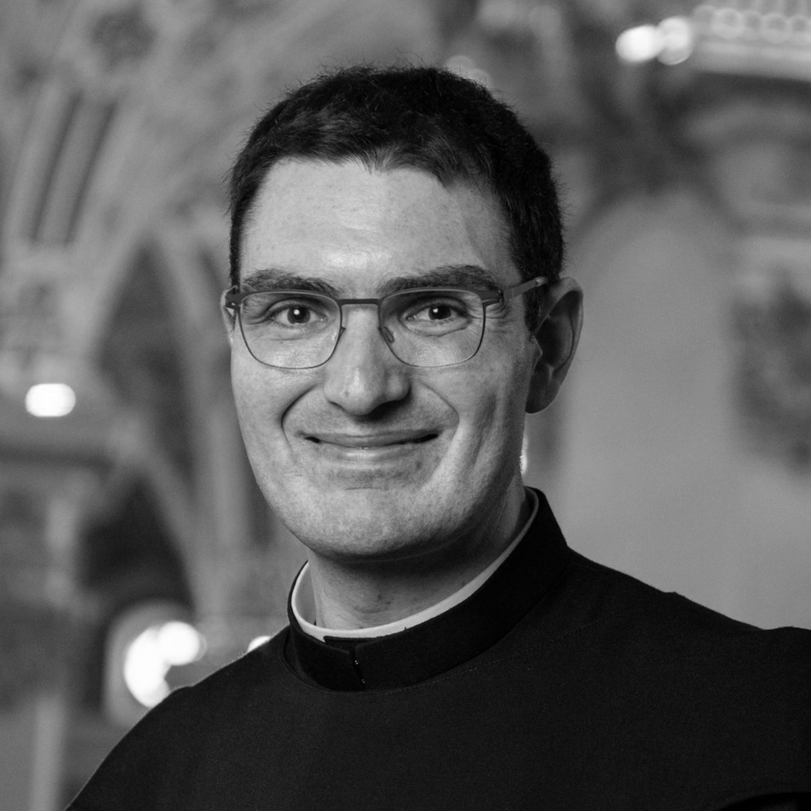 Schwarz-Weiß-Foto eines Mannes mit Brille in einem schwarzen Talar, vermutlich ein Priester, vor einer Backsteinarchitektur im Hintergrund, lächelnd.