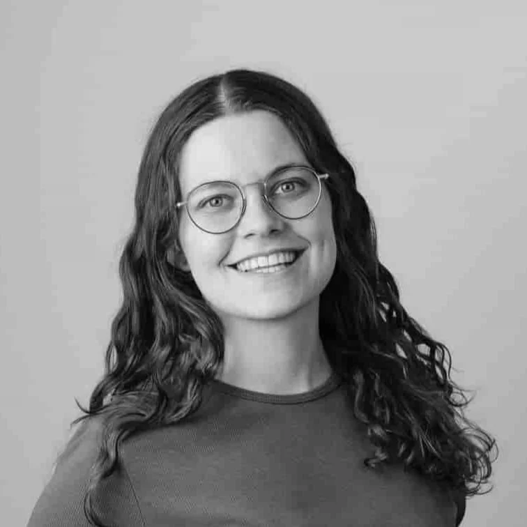 Schwarz-weiß-Porträt einer lächelnden Frau mit Brille und lang, lockigem Haar, gegen einen neutralen Hintergrund.