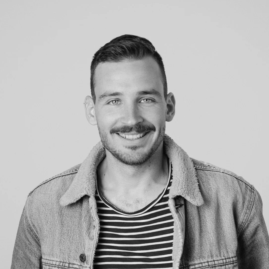 Schwarz-weiß Porträt eines lächelnden jungen Mannes mit kurzem Haar, Bart und Buchstabenmuster-Shirt, in Jeansjacke, vor neutralem Hintergrund.