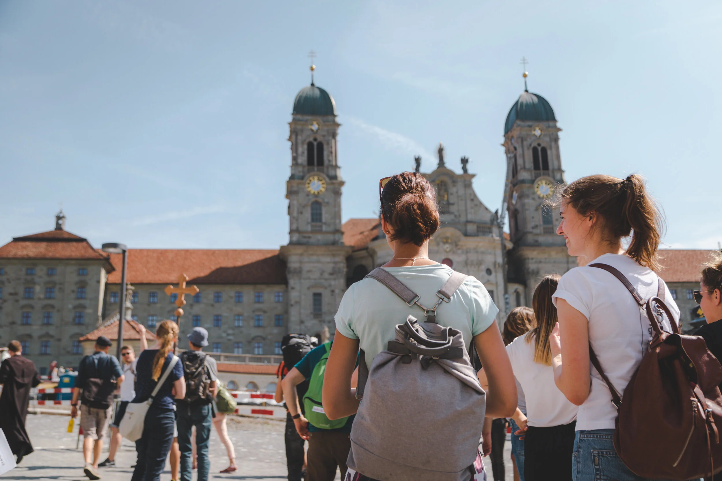 Menschen vor einer barocken Kirche mit zwei Türmen im Hintergrund, im Freien bei sonnigem Wetter.