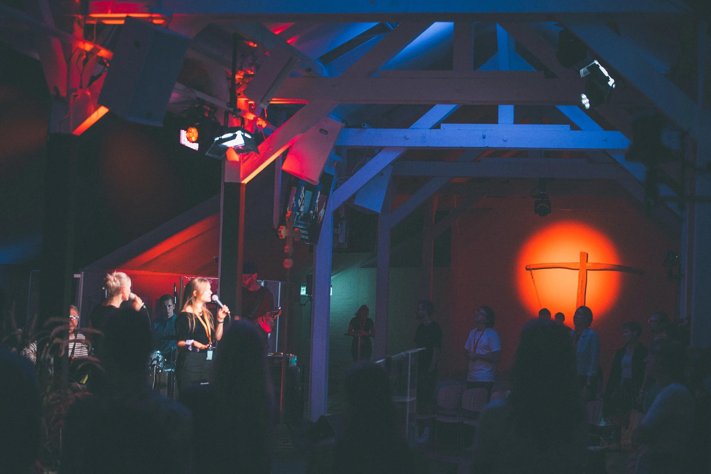 Menschen in einer Kirche mit einem Kreuz im Hintergrund, leuchtturmähnlich beleuchtet, im Rahmen einer Veranstaltung mit Mikrofonen und Bühnenbeleuchtung.
