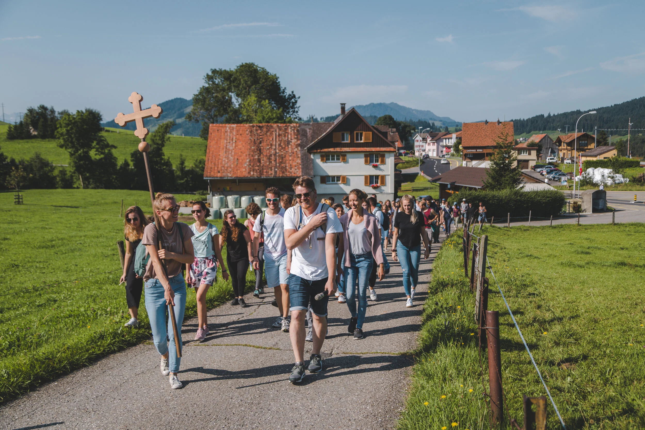 Gruppe von Menschen, die eine ländliche Straße entlanggehen, bei sonnigem Wetter, mit landwirtschaftlichen Gebäuden und grünen Hügeln im Hintergrund.