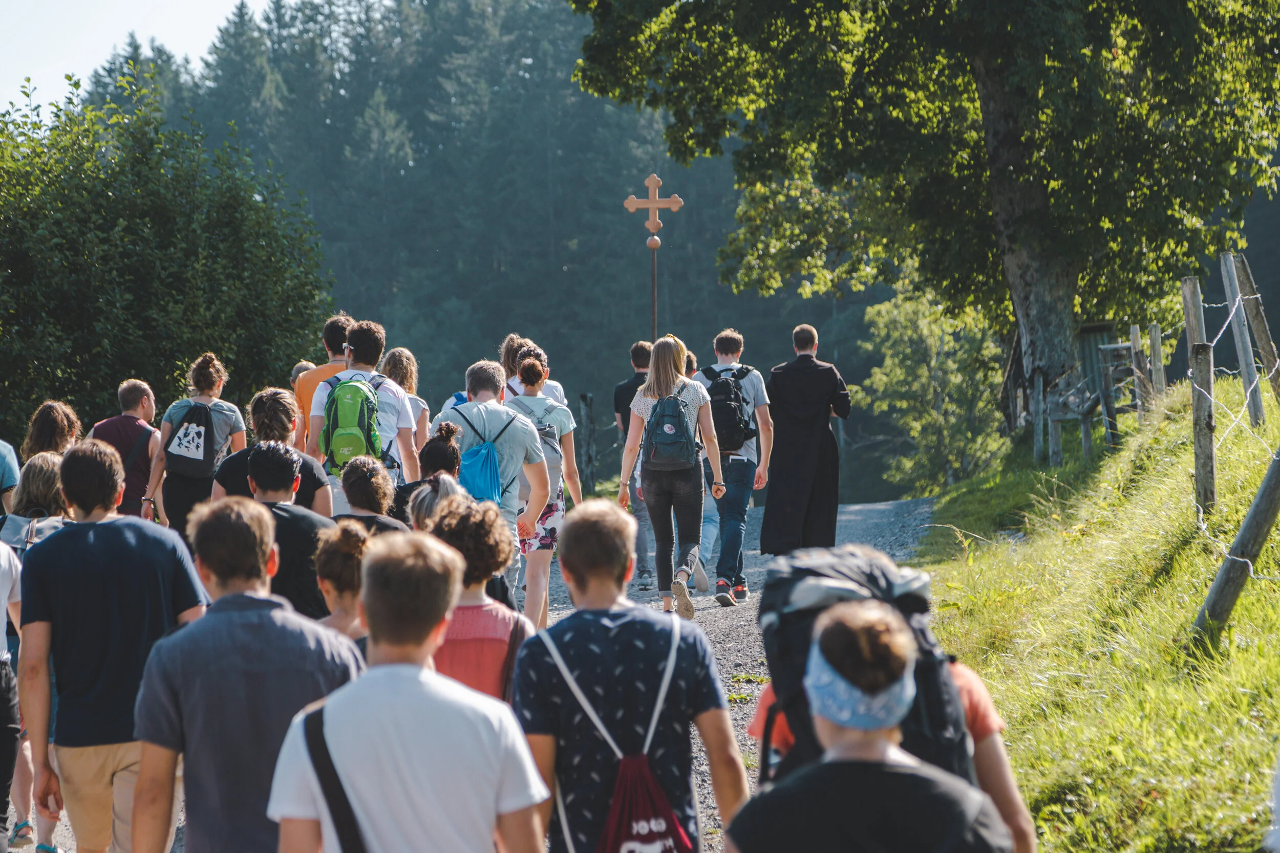 Eine Gruppe von Menschen wandert auf einem unbefestigten Weg in einer ländlichen, grünen Umgebung, mit einem Holzkreuz in der Ferne.