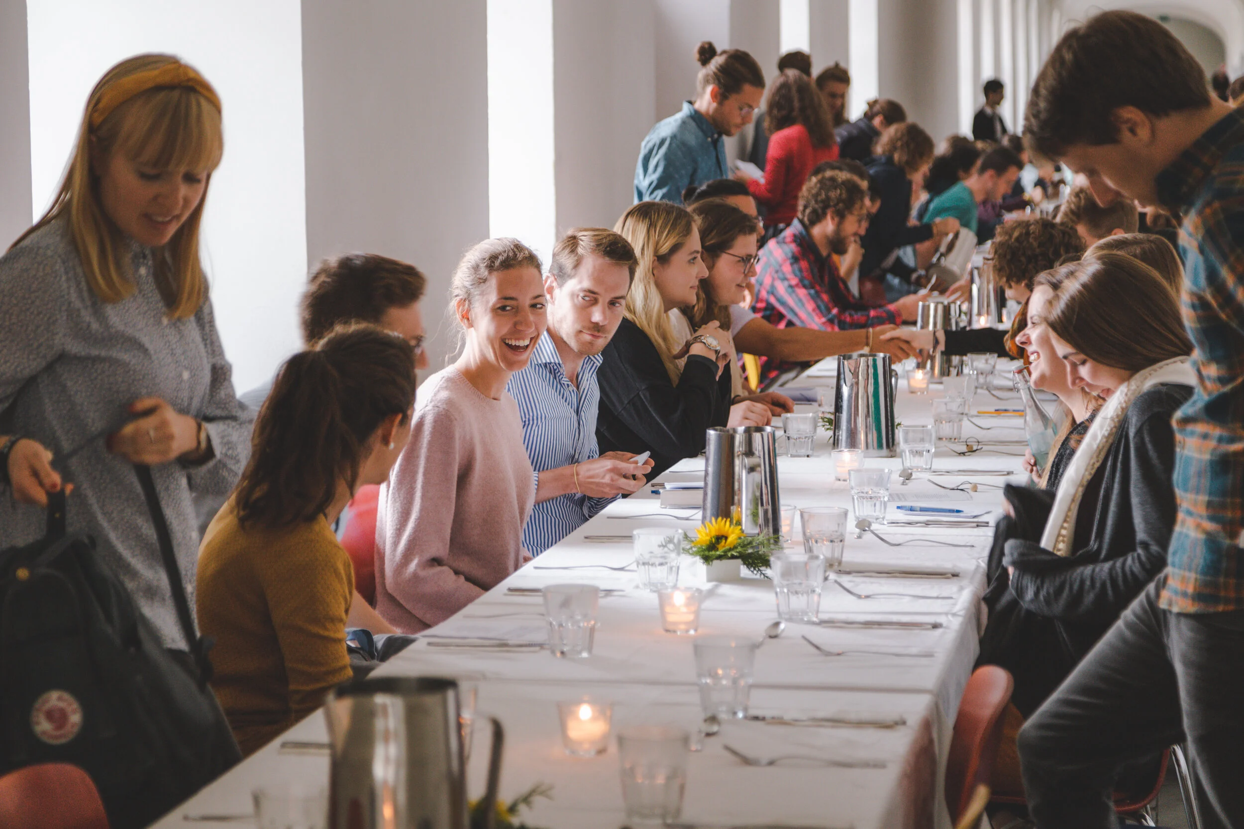 Menschen sitzen an einem langen festlich gedeckten Tisch, einige sind im Gespräch, andere reinigen ihre Hände, im Hintergrund stehen weitere Gäste.