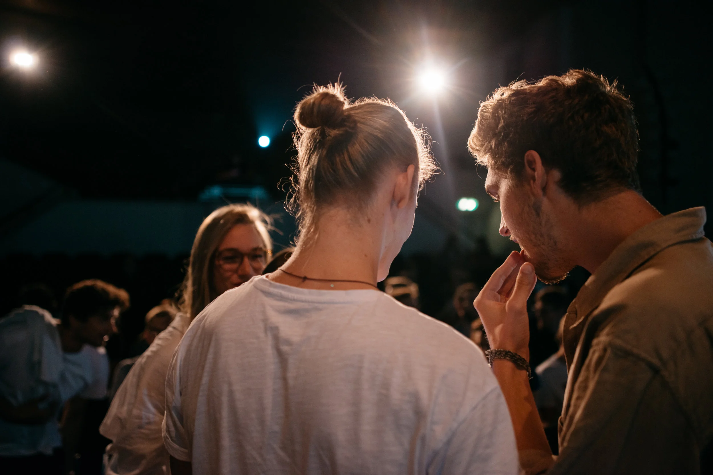 Zwei junge Menschen sprechen eng beieinander in einer dunklen Umgebung mit starkem Licht hinter ihnen, während im Hintergrund weitere Leute sichtbar sind.