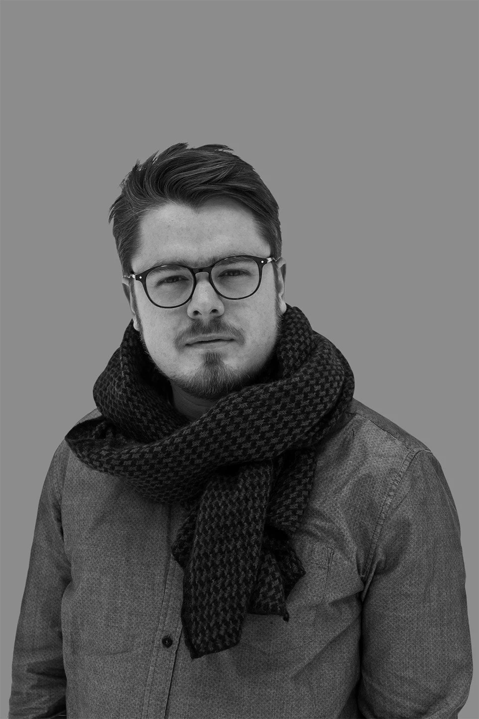 Schwarz-weiß Portrait eines jungen Mannes mit dunklem Haar, Brille und Bart, der eine dunkle Jacke und einen gehäkelten Schal trägt