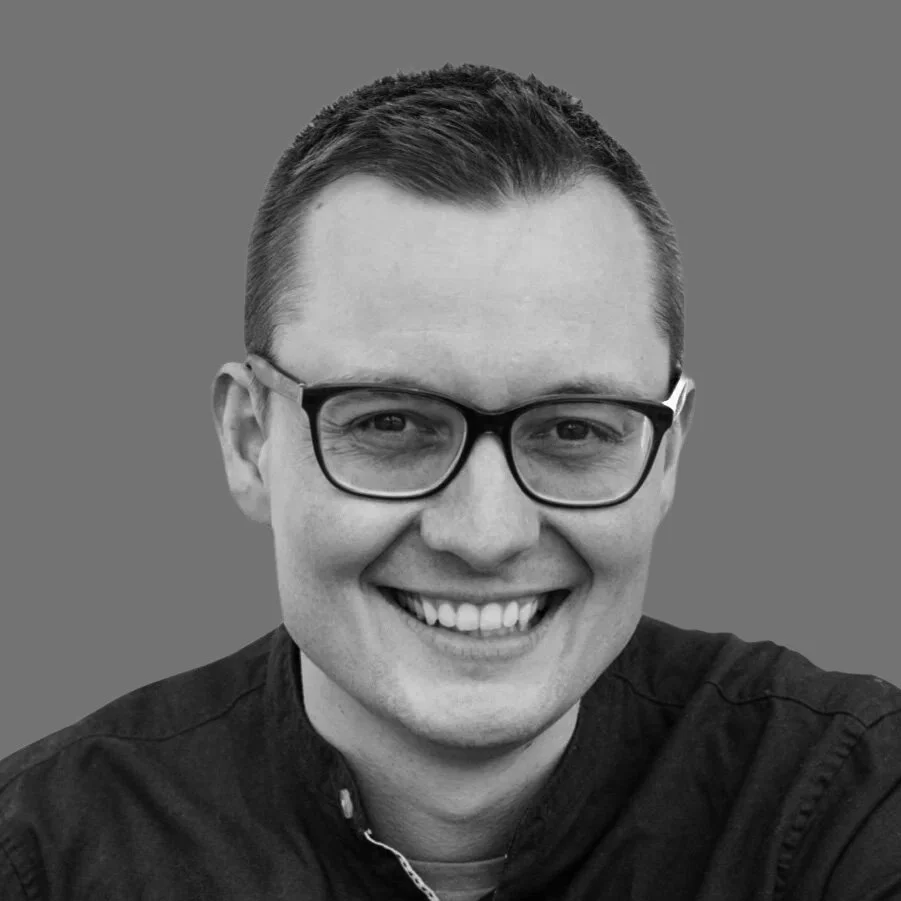 Schwarz-weiß-Foto eines lächelnden Mannes mit Brille und kurzen Haaren vor einem grauen Hintergrund.