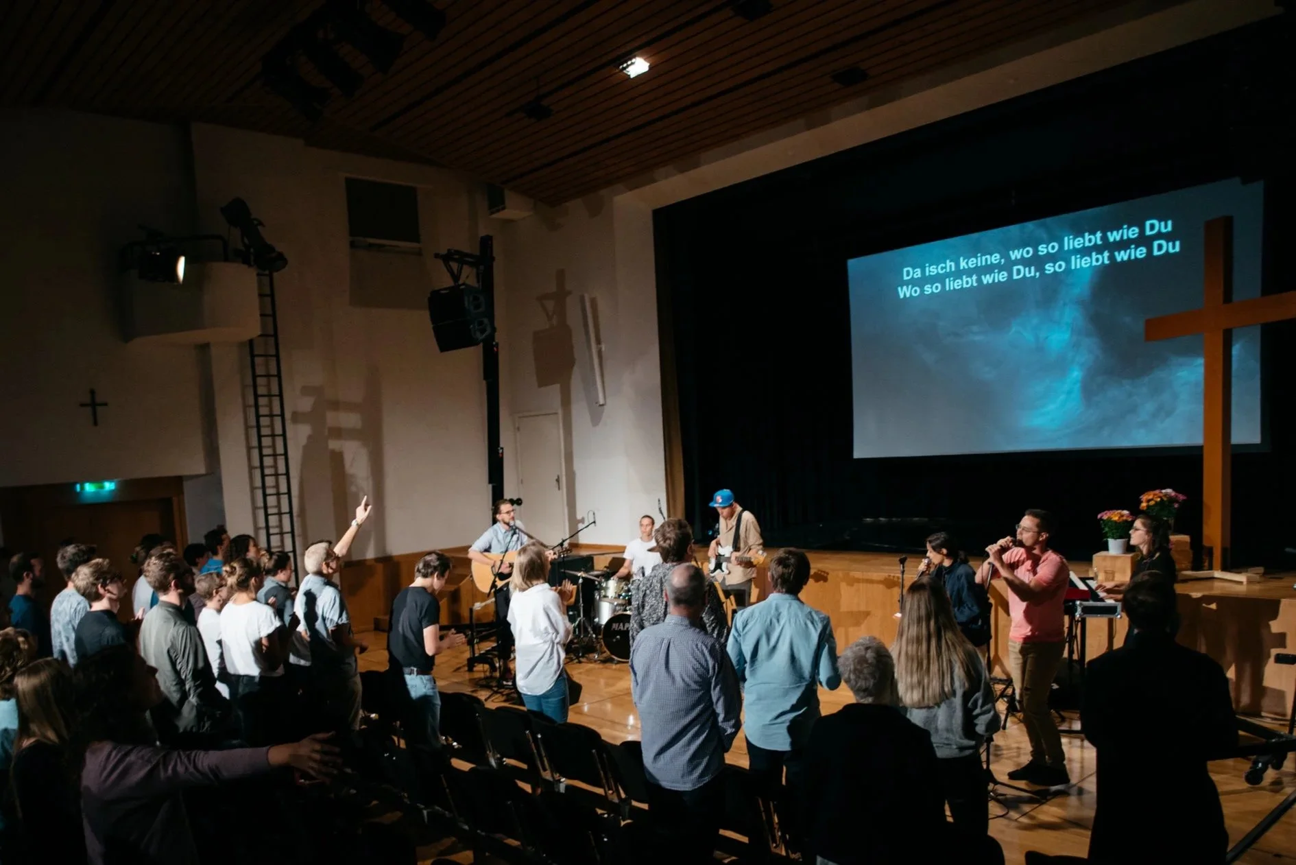 Menschen in einer Kirche beim Singen und Musizieren, während auf der großen Leinwand Liedtexte angezeigt werden, mit einem Kreuz und Blumen auf der Bühne.