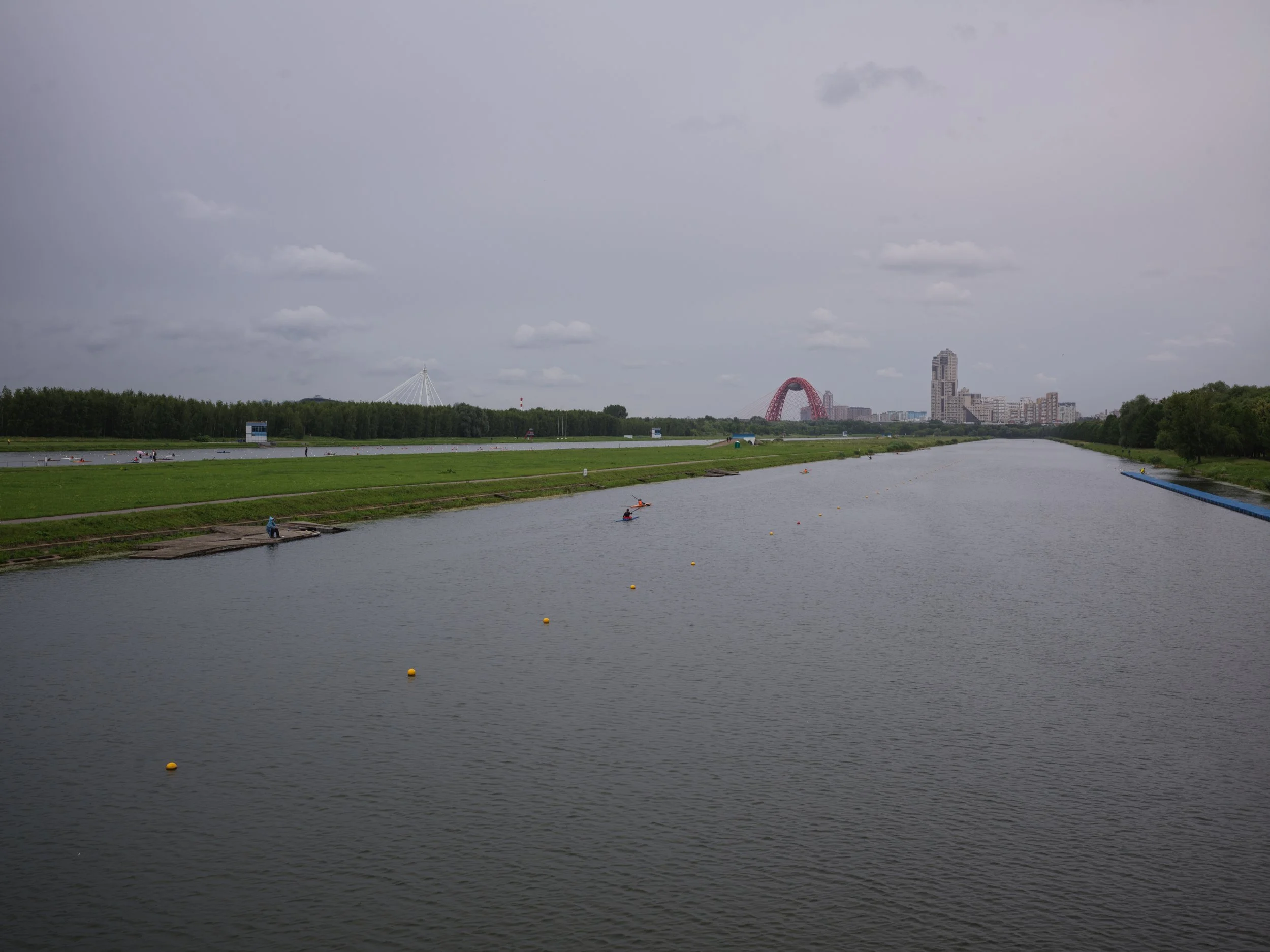 A wide view of a river with a city skyline in the background, cloudy sky, and a few people kayaking on the water.