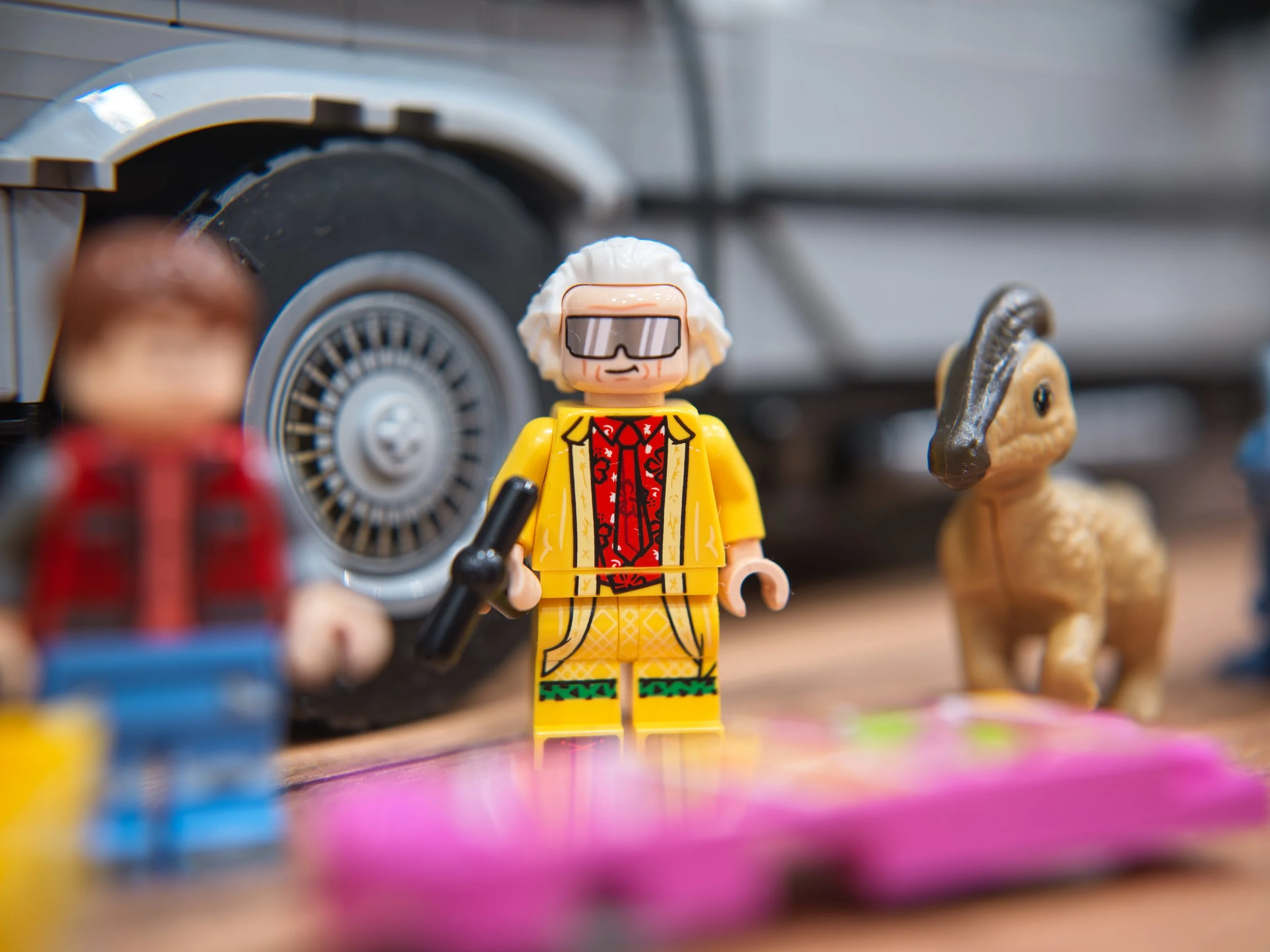 Lego figure of a man with white hair wearing sunglasses, a yellow jacket, and a red shirt, holding a black object, standing on a pink surface with toys and a car in the background.