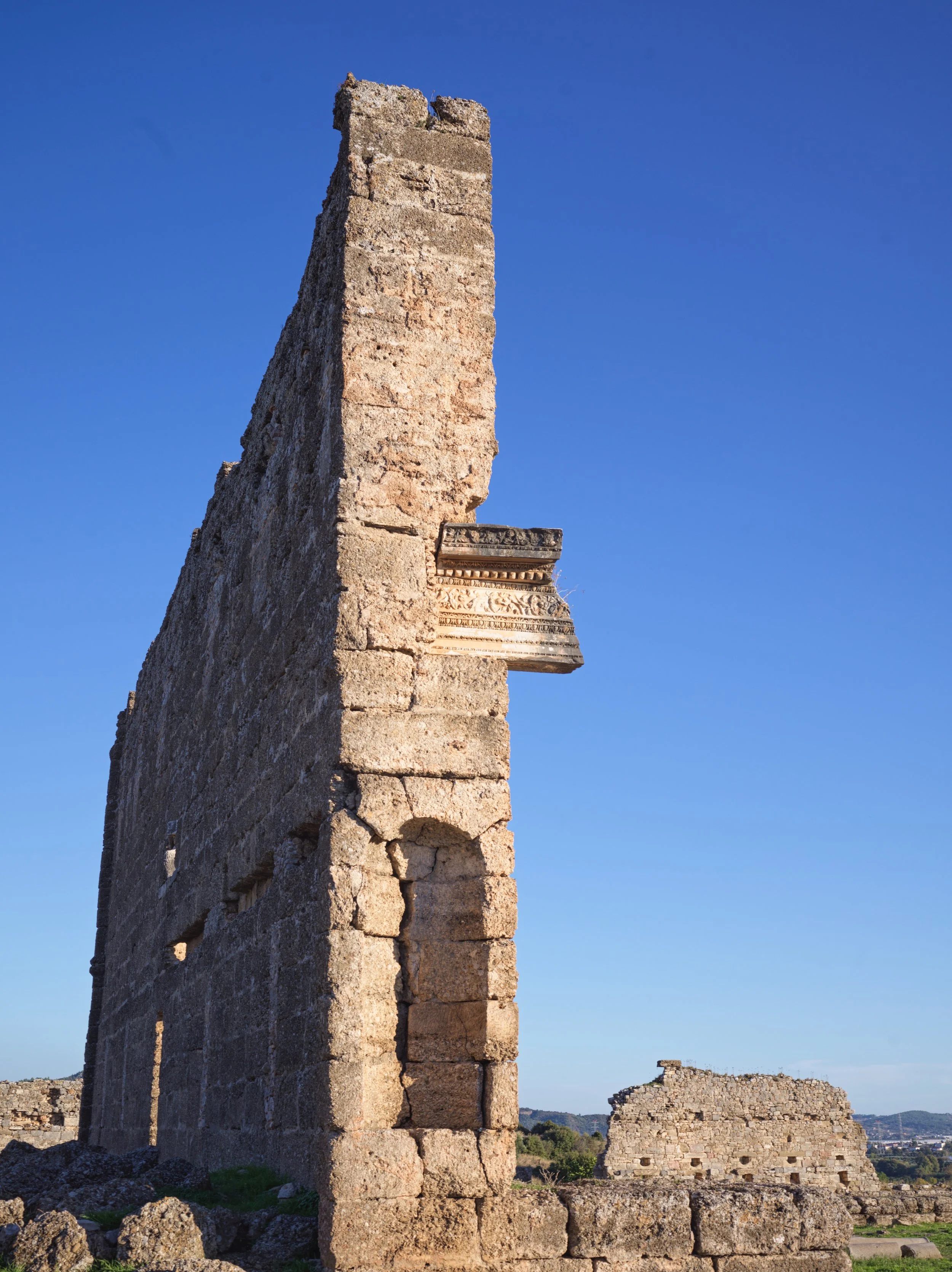 Ancient stone ruin with a tall, weathered wall and decorative frieze, under a clear blue sky.