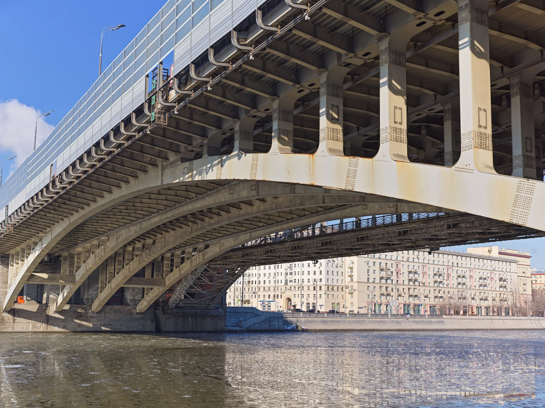 A large steel bridge spanning over a river in an urban area, with the city skyline and buildings in the background under a clear blue sky. Photo by Alexander Sokolyuk. Moscow, Russia