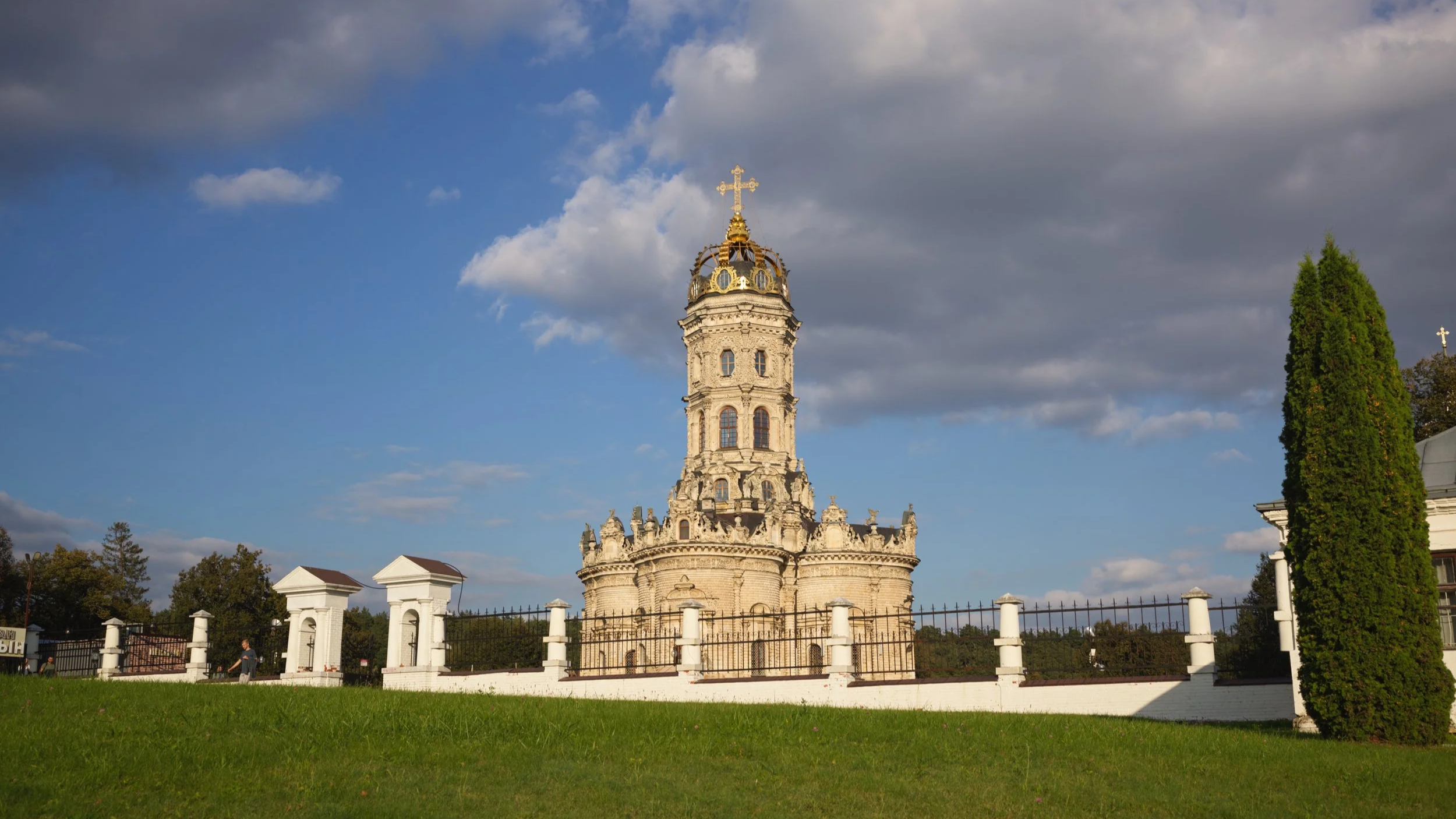 A historic church with a tall ornate bell tower is surrounded by a white fence and lush green grass. The sky above is partly cloudy with patches of blue.
