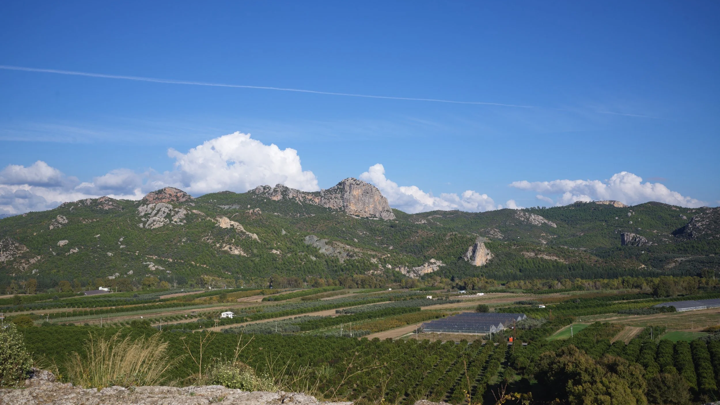 Scenic landscape of a mountain range with green hills, fields, and farm buildings under a blue sky with white clouds.