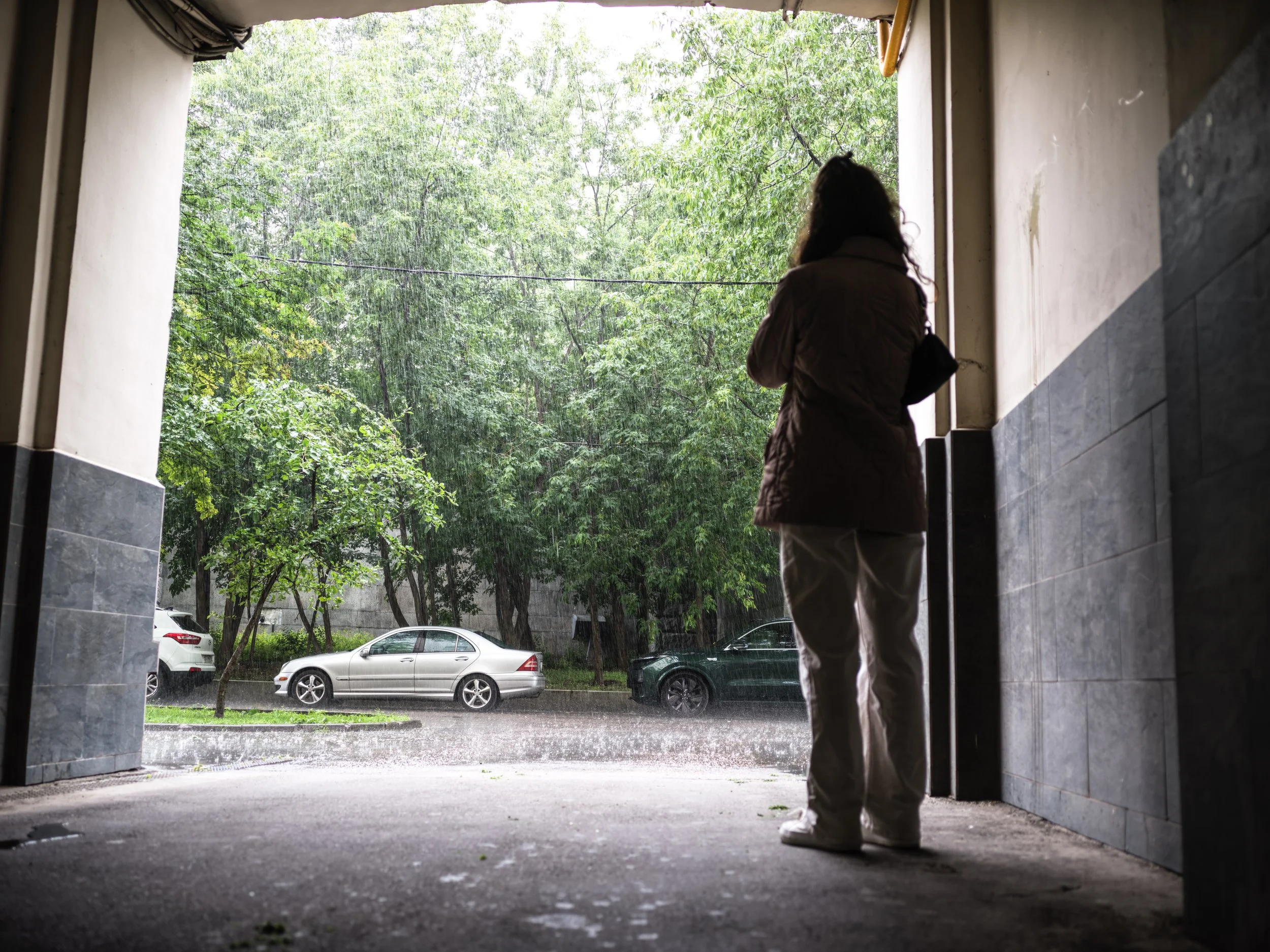 Person standing inside a parking garage, looking out at a rainy street with trees and parked cars.
